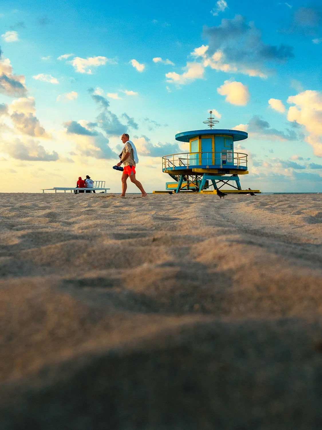 Man walking barefoot on South Beach sand past a blue and yellow Art Deco lifeguard stand at golden hour