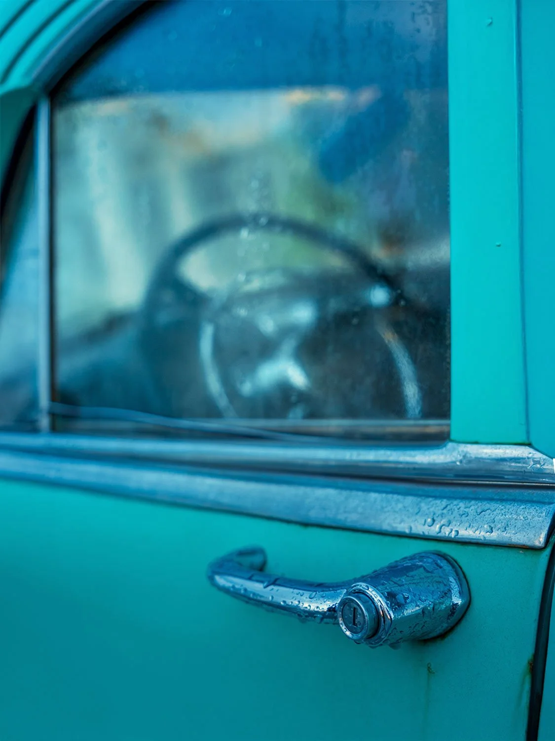 Close-up of a turquoise vintage car door with chrome handle and rain droplets, steering wheel visible through window
