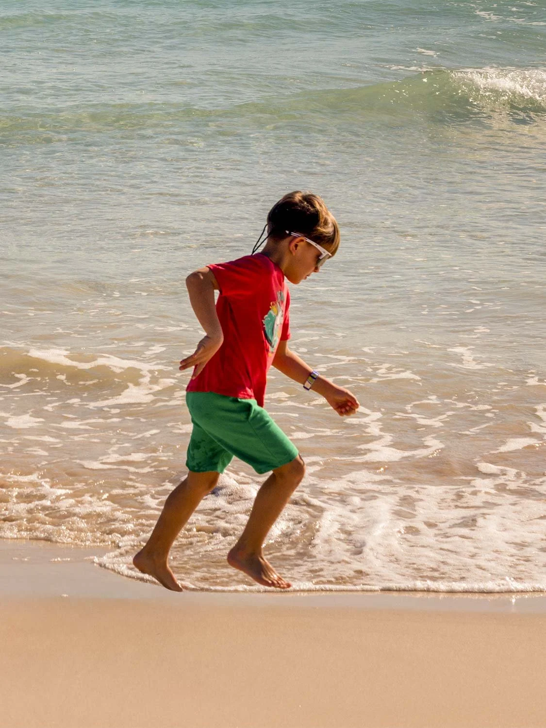 Child in red shirt and green shorts running barefoot along sandy beach shoreline with ocean waves