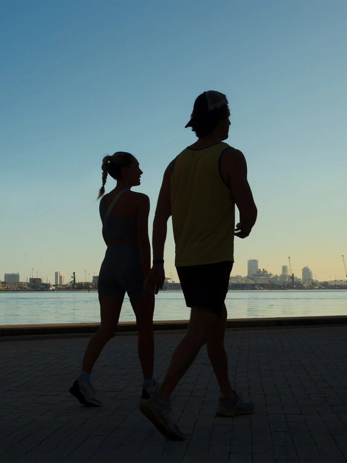 Couple in workout clothes jogging along urban waterfront at sunset, wearing athletic shorts, sports bra, and tank top.