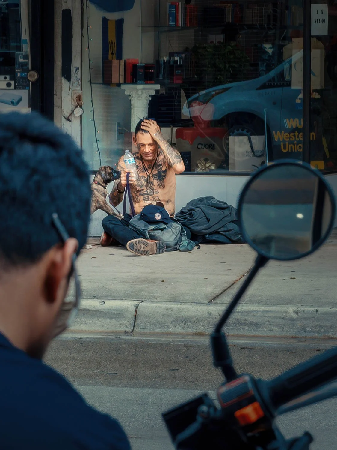 Shirtless tattooed man sitting on a Miami sidewalk with his dog, drinking water outside a storefront, framed by a passerby and motorcycle mirror