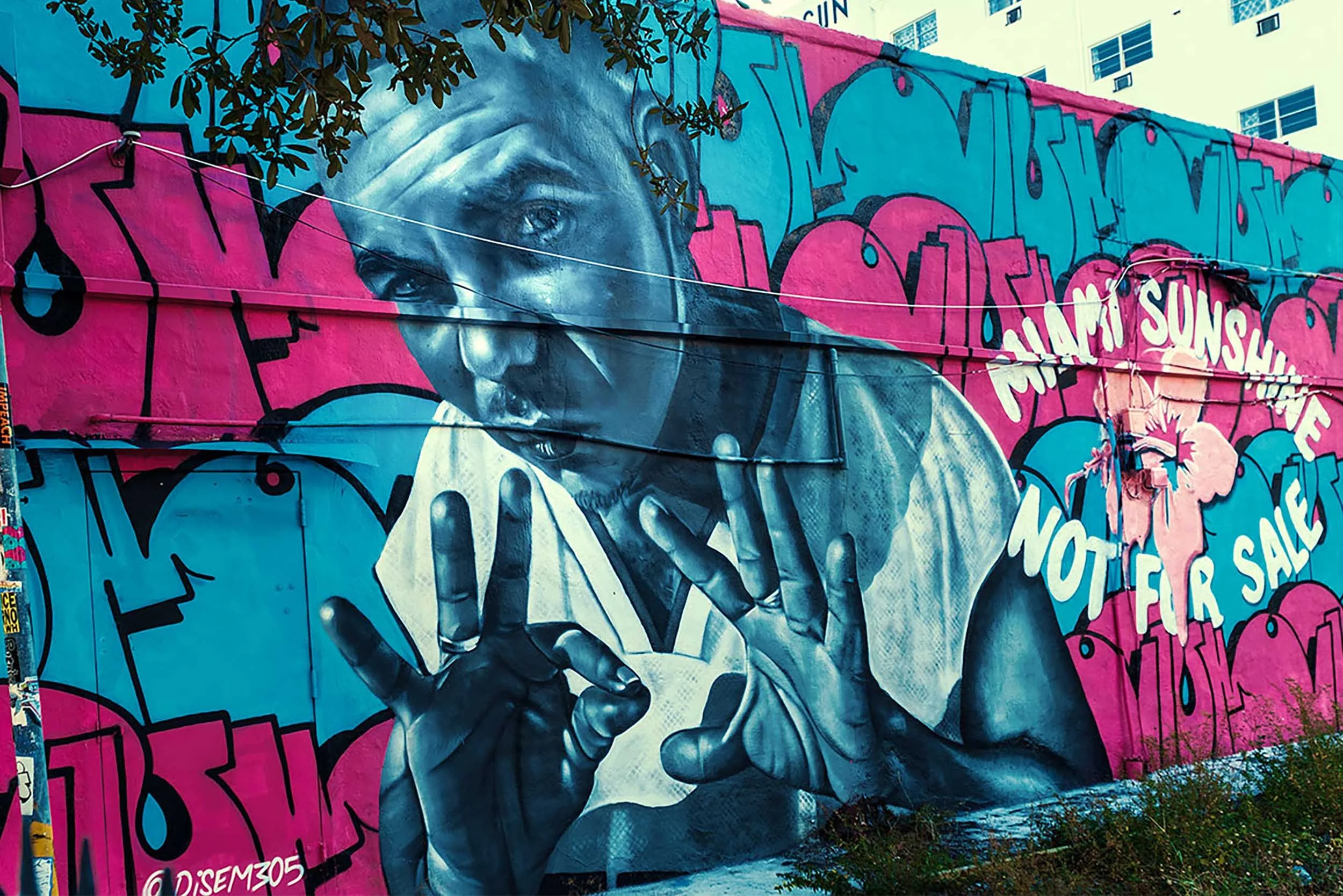 A black-and-white portrait of a man wiping his face with a towel over pink and blue lettering, signed @DISEM305