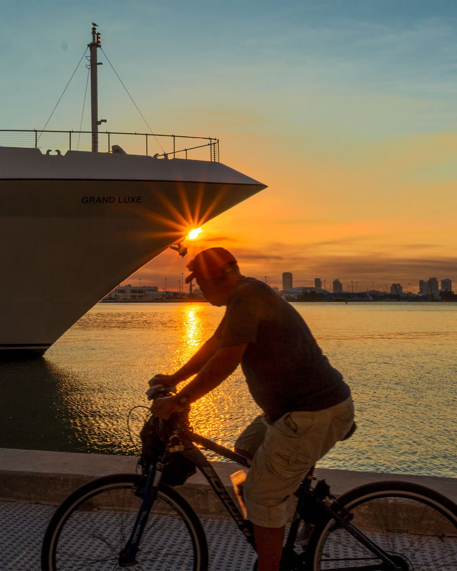 Cyclist riding past a docked cruise ship bow with a sun flare over the bay, Bayfront Park Miami