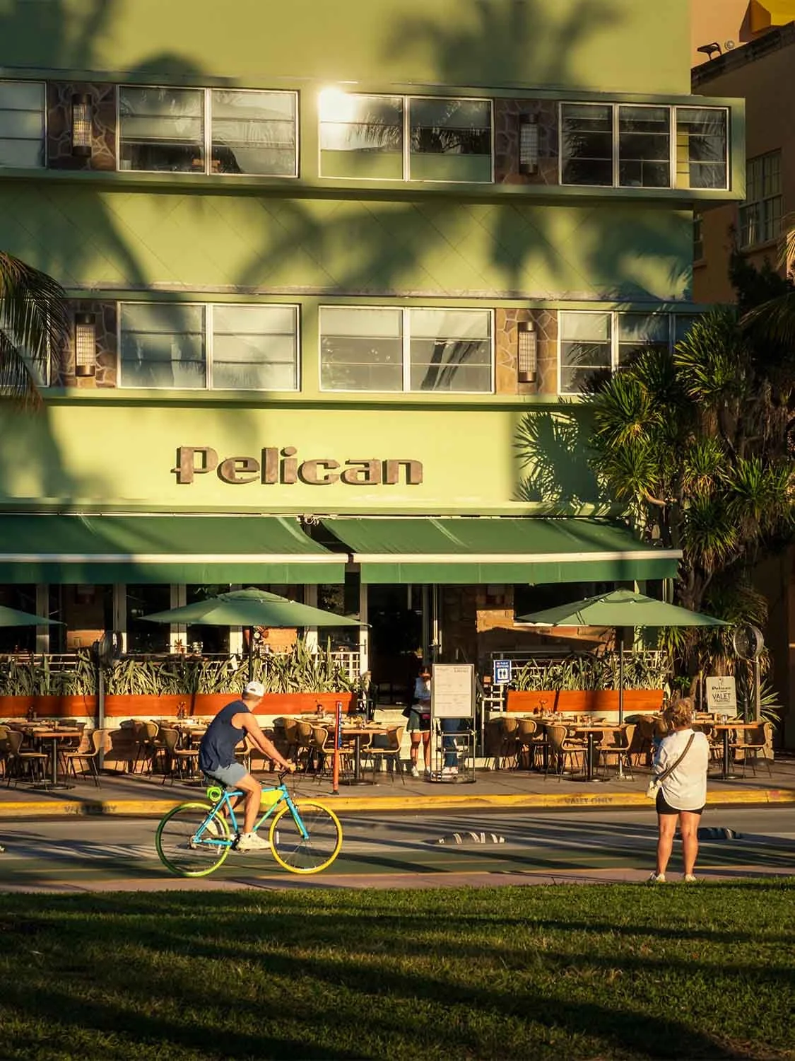 Cyclist rides past Pelican Hotel outdoor patio with green awning and umbrella seating on Miami Beach