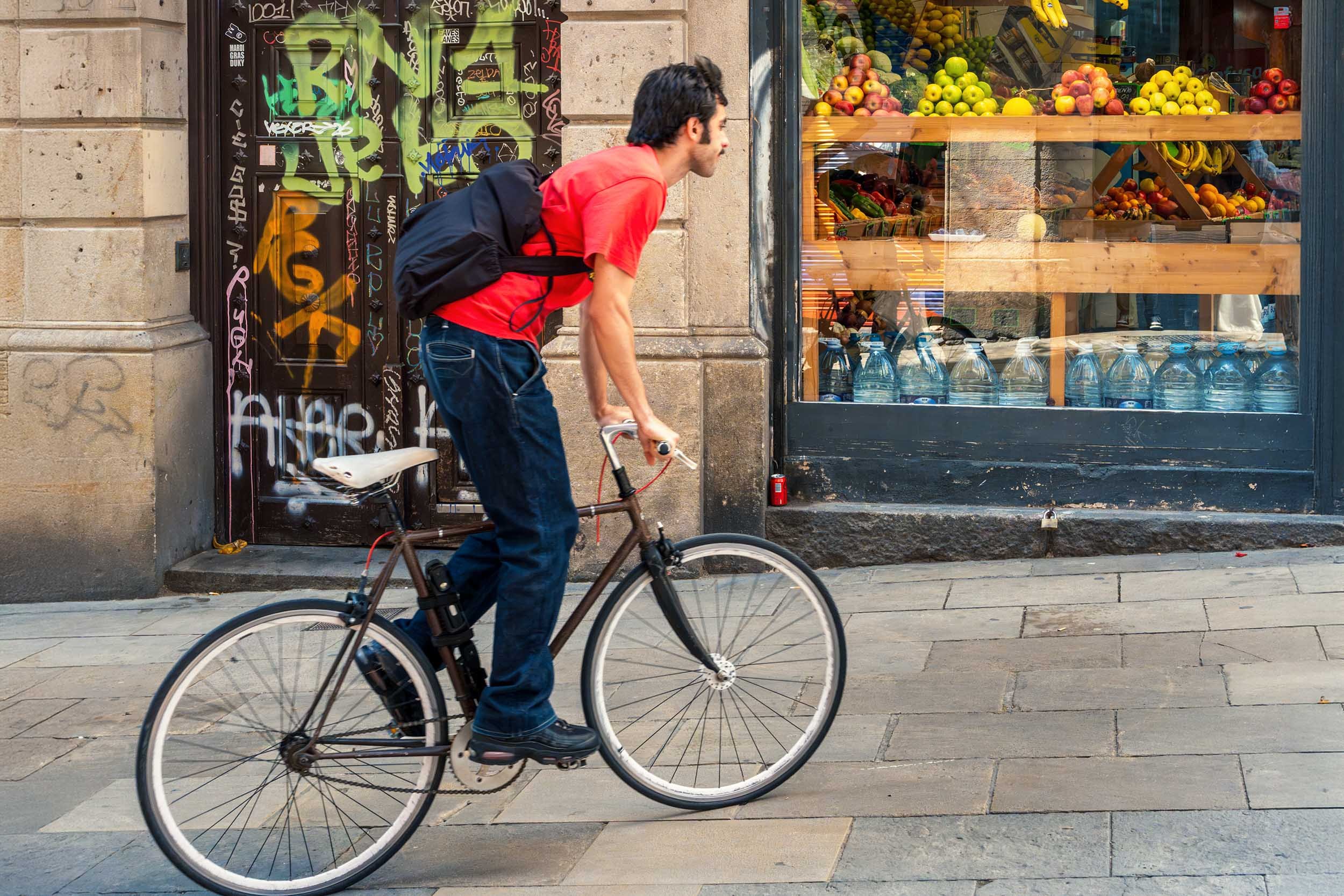 Cyclist in a red t-shirt riding past a graffiti-covered wooden door and a fruit shop window on a stone-paved street