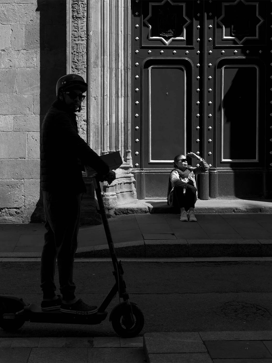 Man on an electric scooter in shadow as a woman shades her eyes on the step of a studded church door in the Gothic Quarter, in black and white
