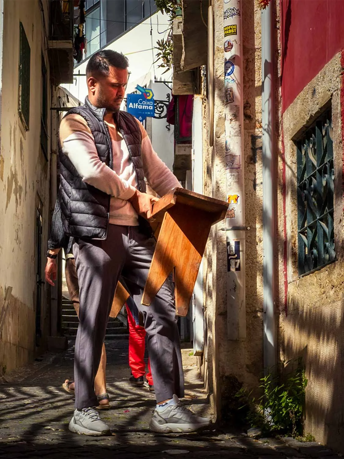 Man carrying a wooden stool down a narrow sunlit alley in Alfama, Lisbon