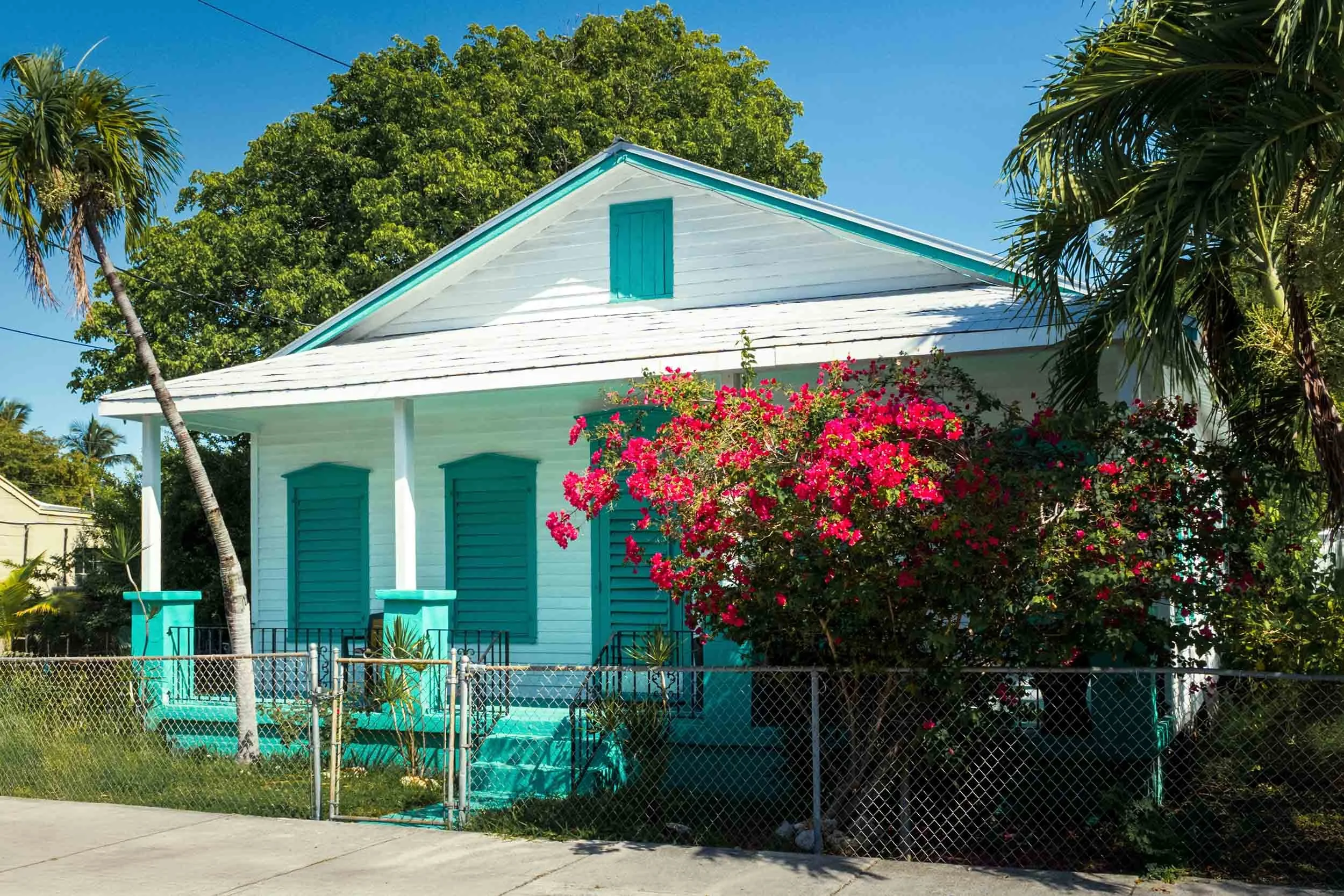 Charming turquoise Key West cottage with vibrant pink bougainvillea, palm trees, and a chain-link fence under blue skies