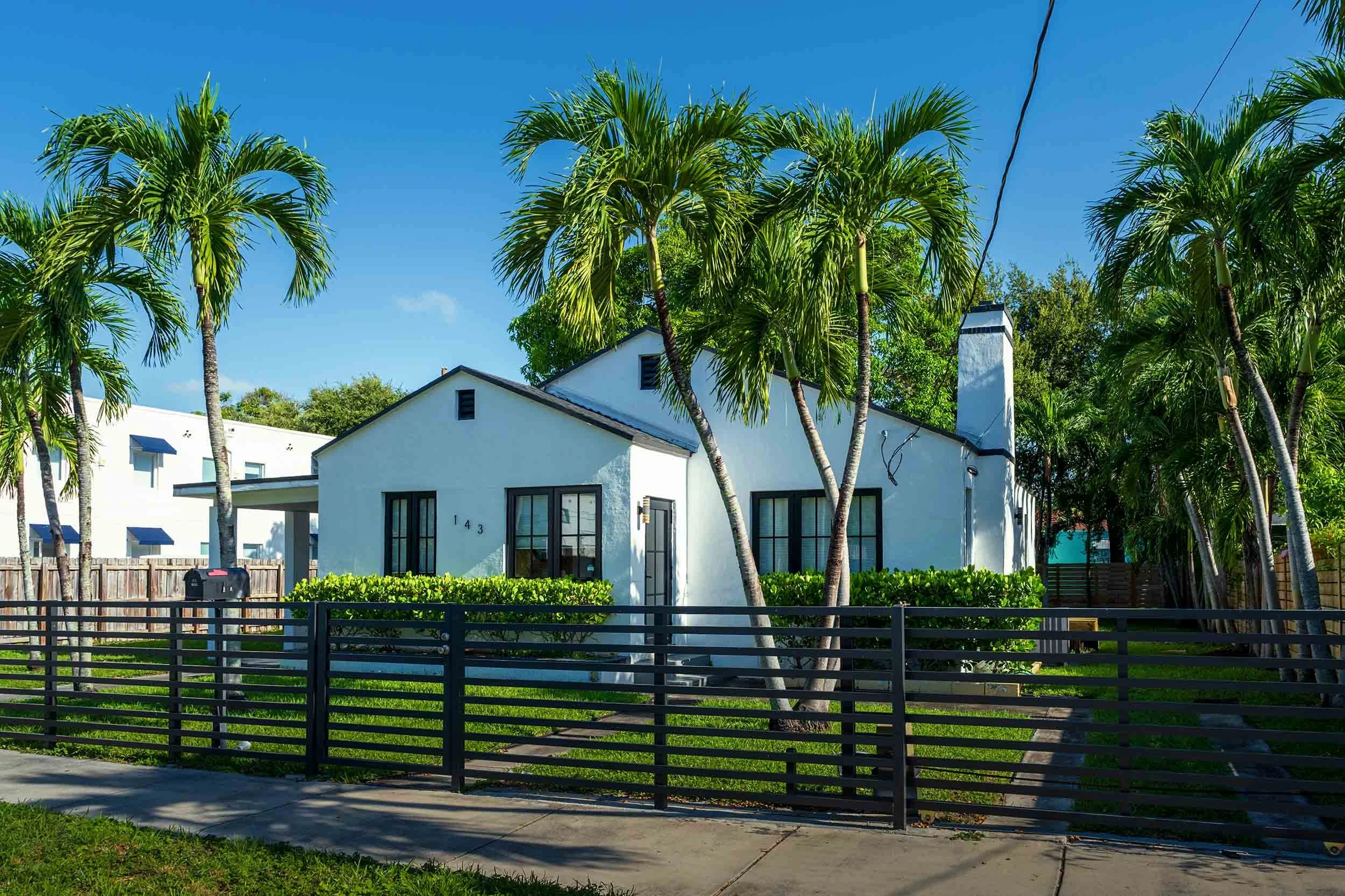 White bungalow with tall palm trees and black slat fence, Buena Vista Miami