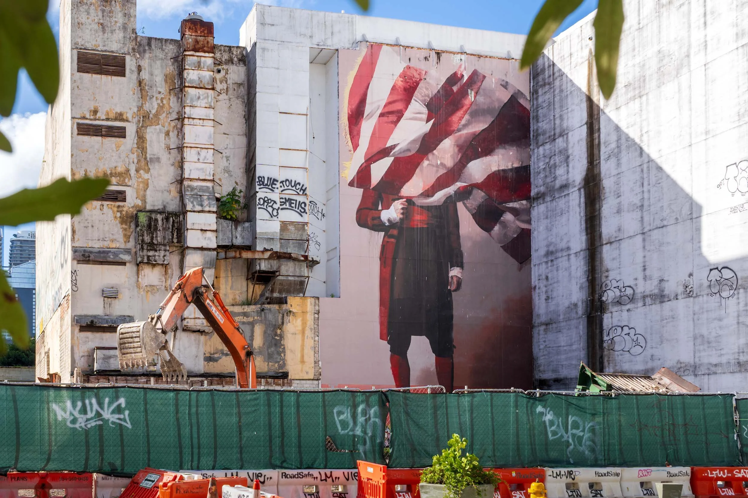 A wide shot of the flag-figure mural and the Fila mural on the same block, with an excavator and construction screening in front