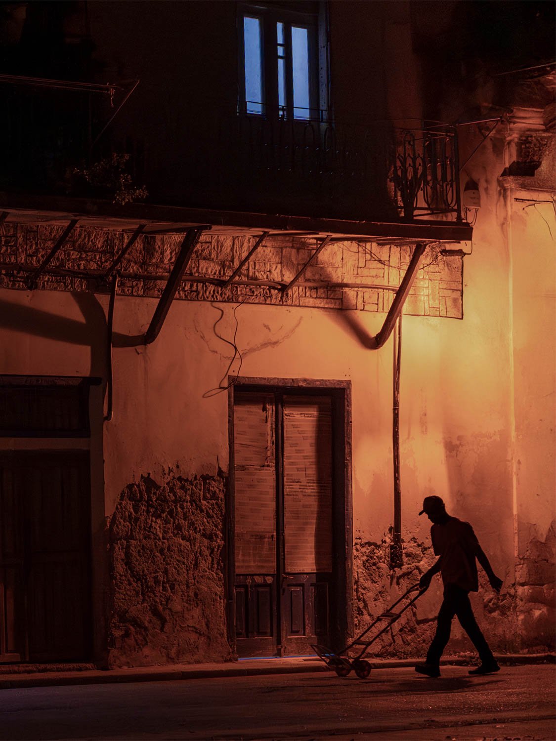 Man pulling a hand cart past a crumbling wall under orange streetlight in Old Havana at night