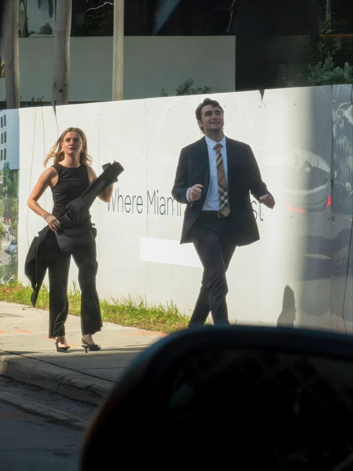 Two people in formal attire walking past a construction hoarding in Miami, photographed through a car window in bright midday light