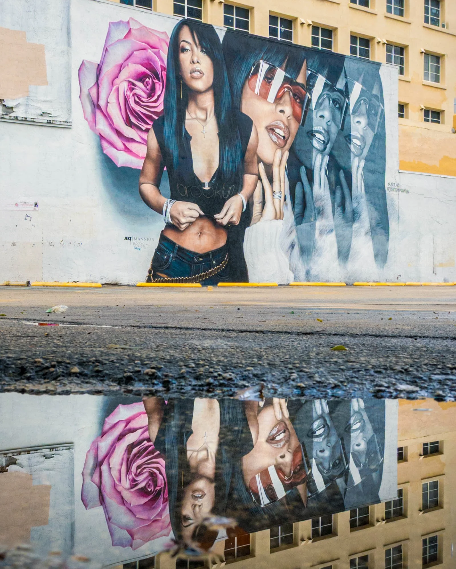A low-angle puddle reflection of the Aaliyah mural, three images of her beside a pink rose, signed JEKS | MANNION