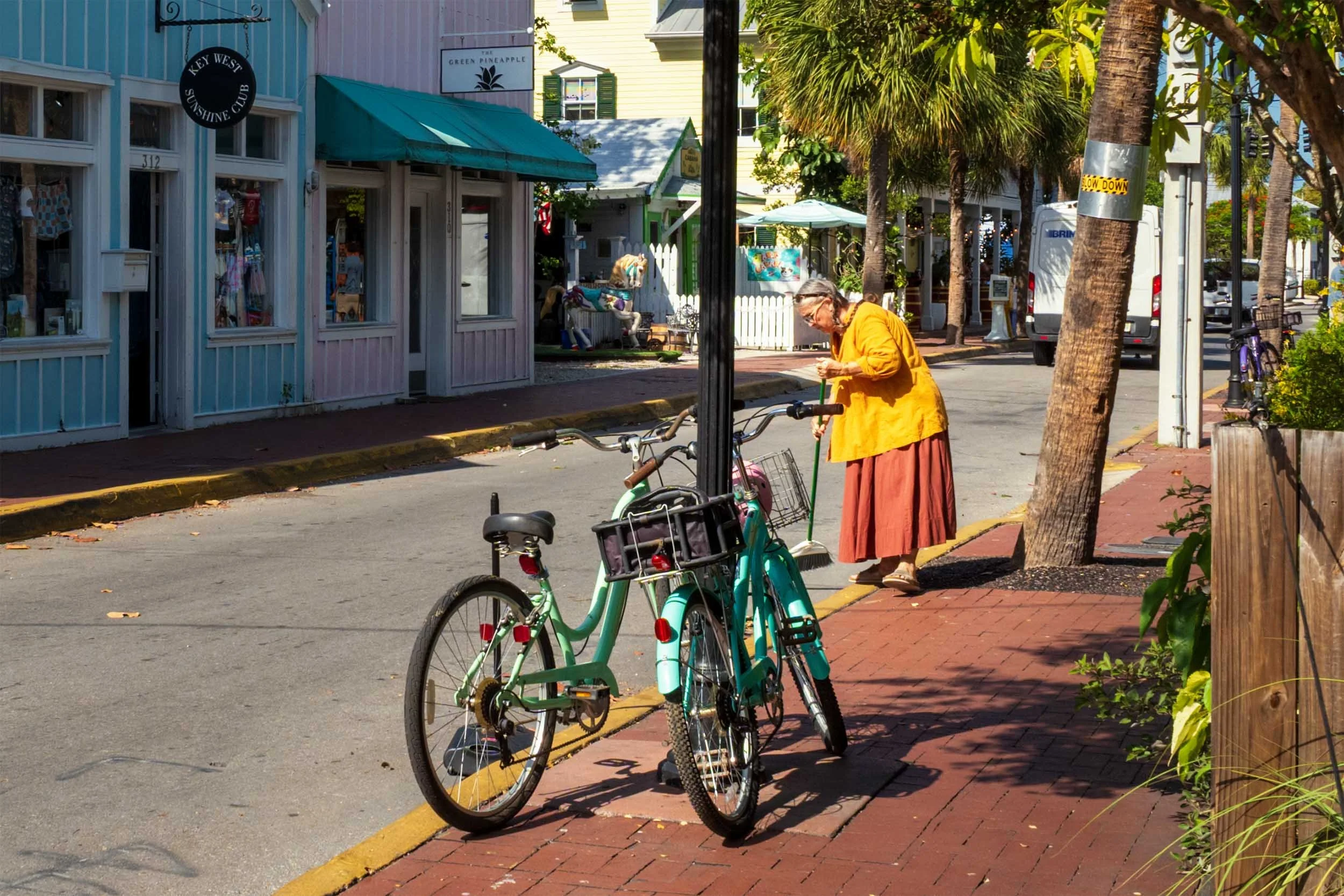 Elderly woman in orange blouse sweeping a brick sidewalk beside teal bicycles on a sunny Key West street