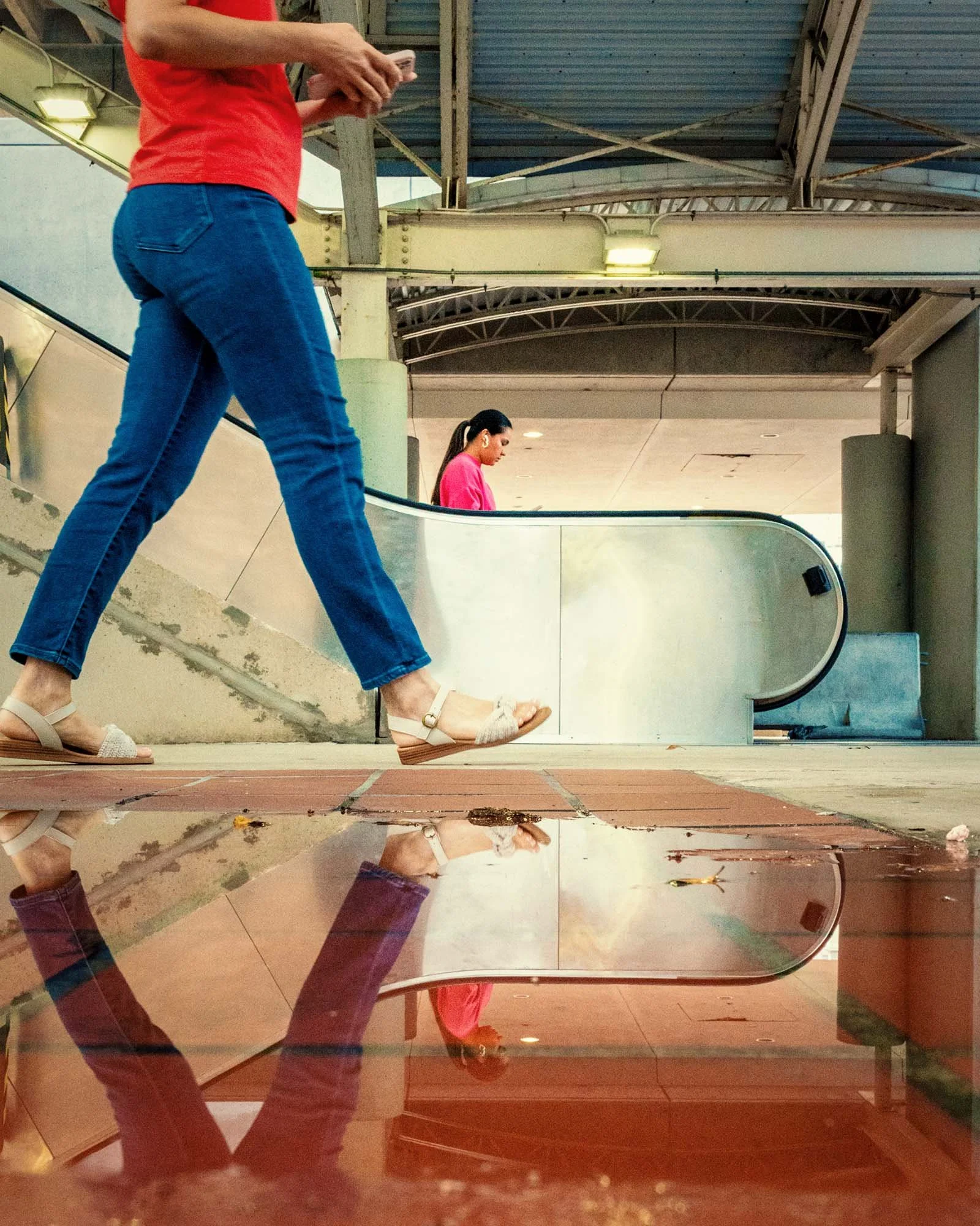 Low-angle street photo of a woman in a red top and jeans walking past an escalator at Government Center station in Miami, her legs reflected in a puddle, with a woman in pink riding the escalator behind her.