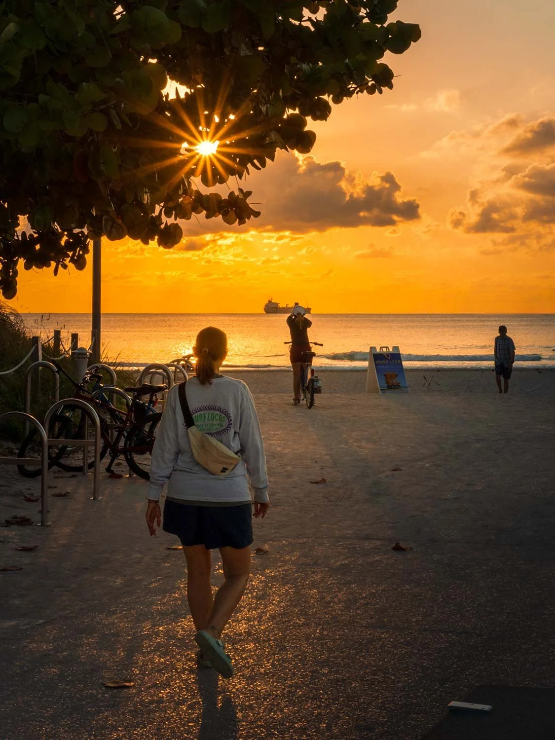 Woman walking toward sunrise at South Beach with golden sky, ocean view, bicycles, and a ship on the horizon