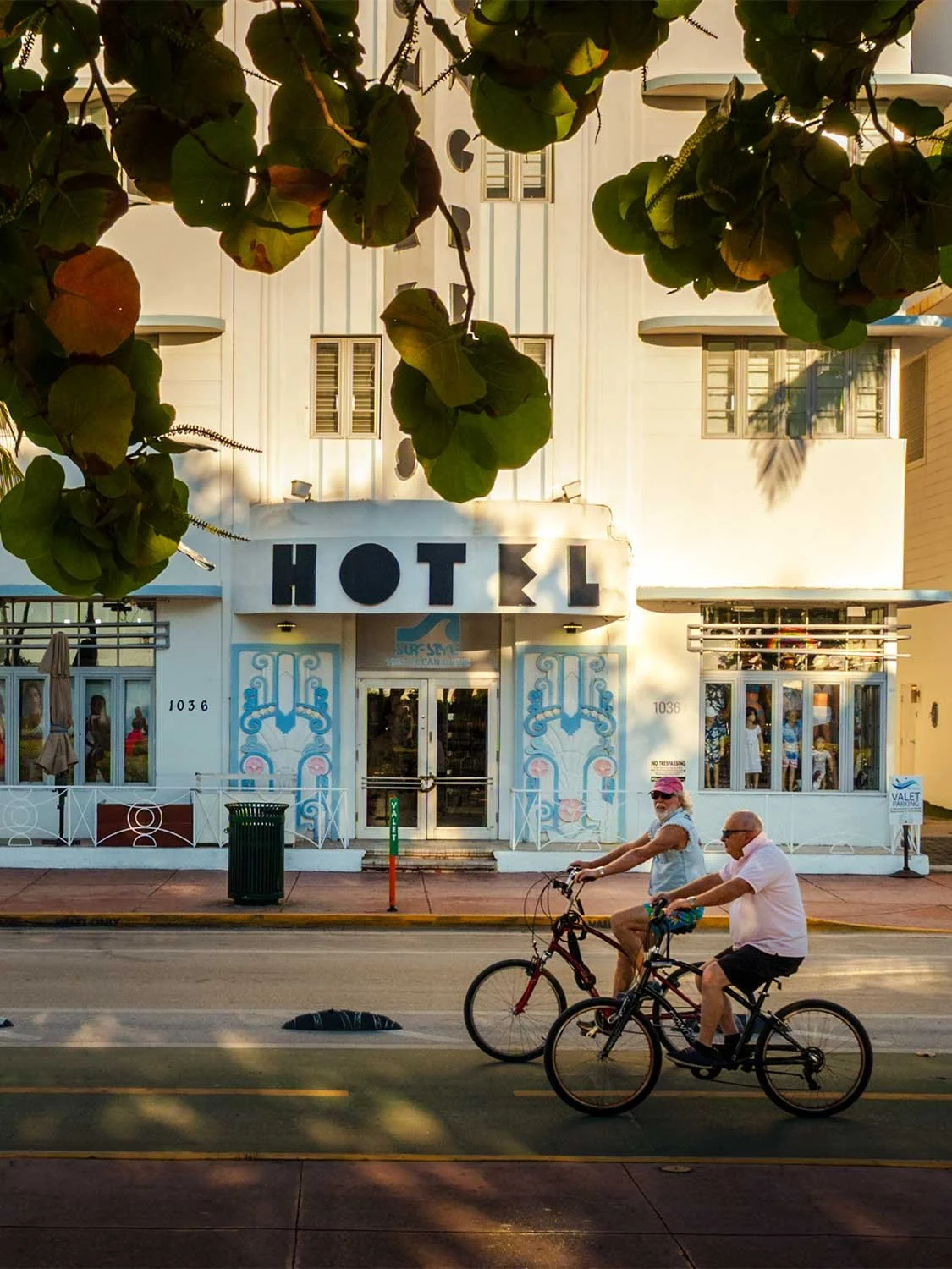 Two cyclists ride past a white Art Deco hotel facade at 1036 Ocean Drive in Miami Beach at golden hour