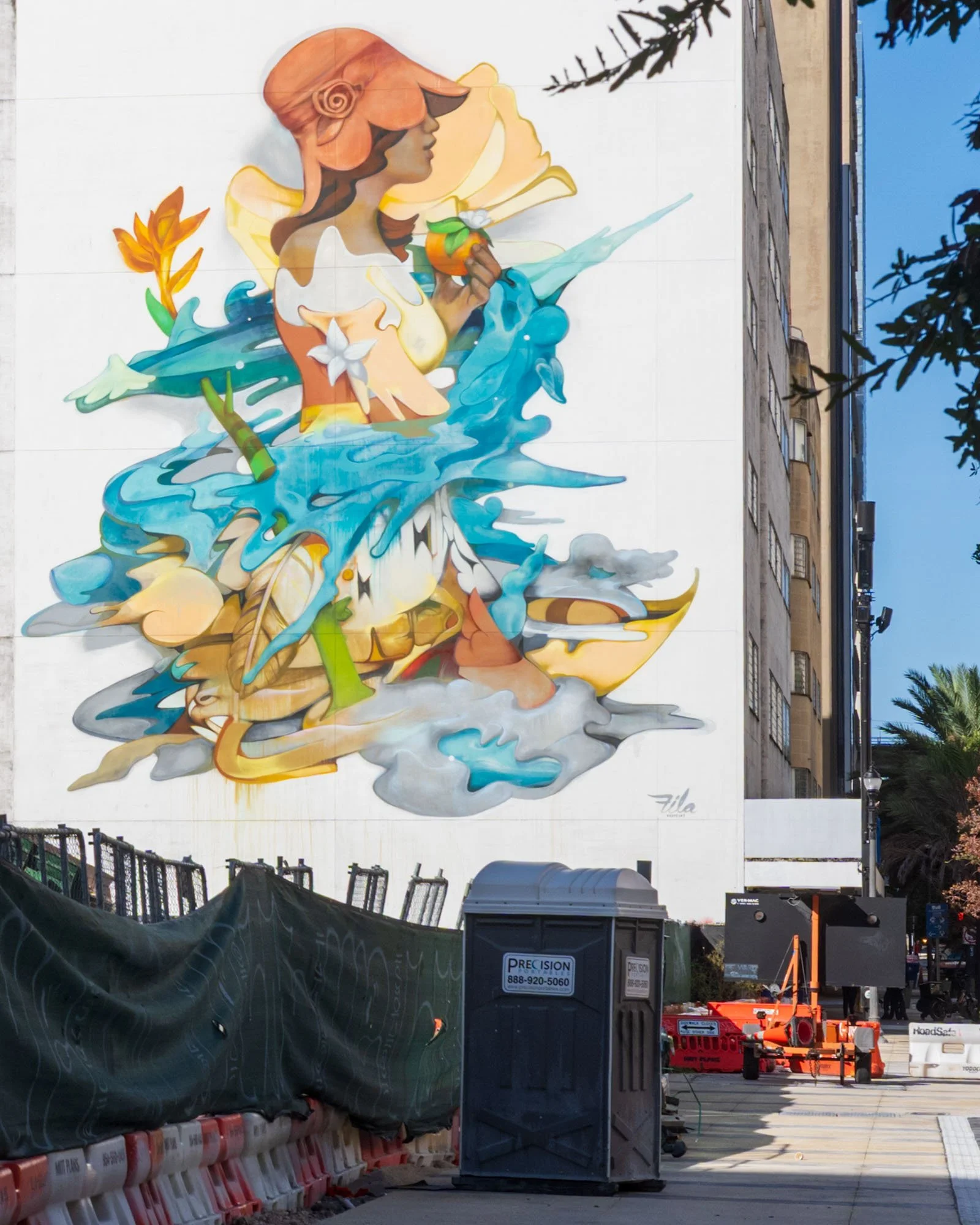 A woman in an orange hat holding an orange, surrounded by blue water shapes and signed Fila, with a construction fence and a portable toilet in front