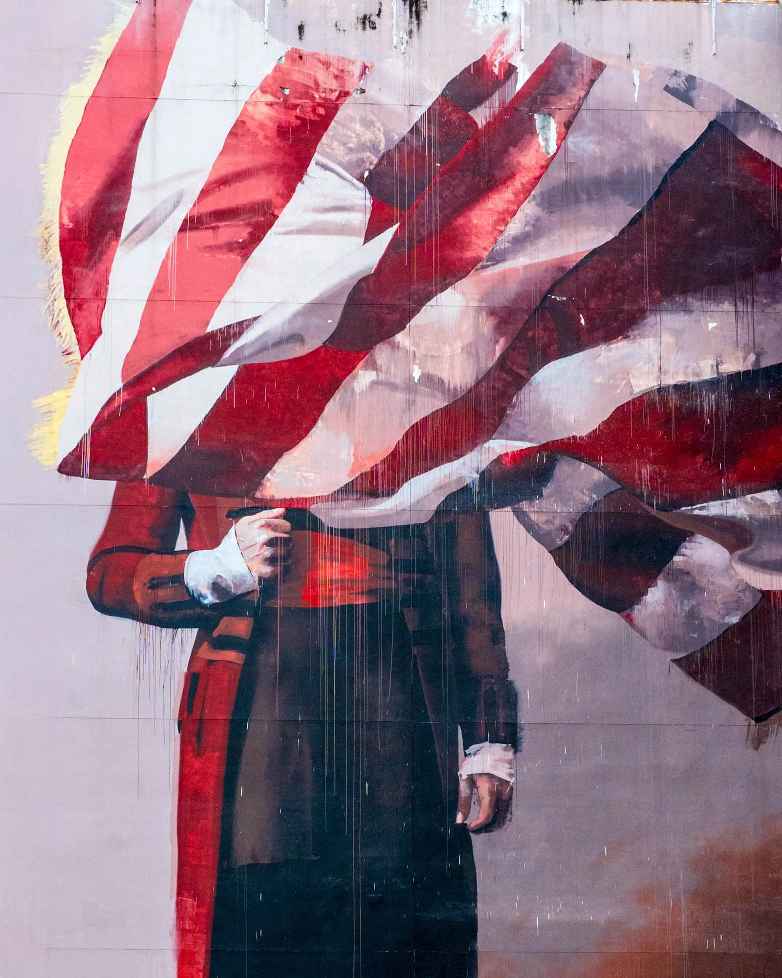 A tight crop of a figure in a red coat with his head covered by a striped flag, and paint drips running down the wall