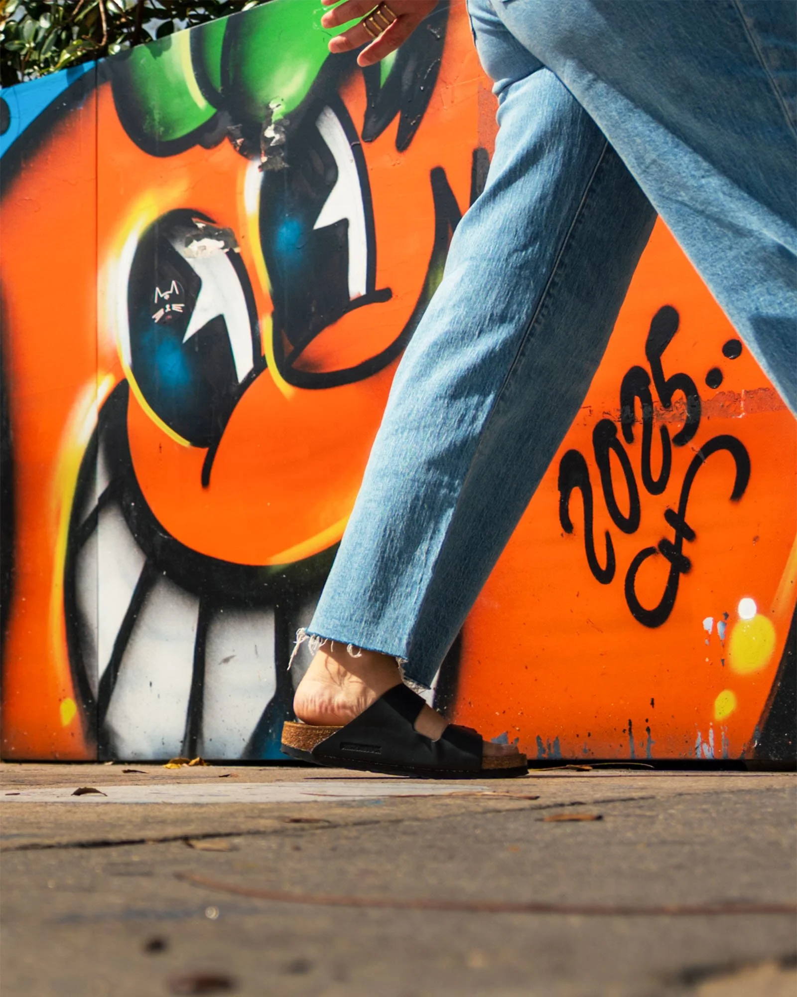 Ground-level close-up of frayed jeans and black sandals mid-stride in front of a bright orange cartoon mural in Wynwood.