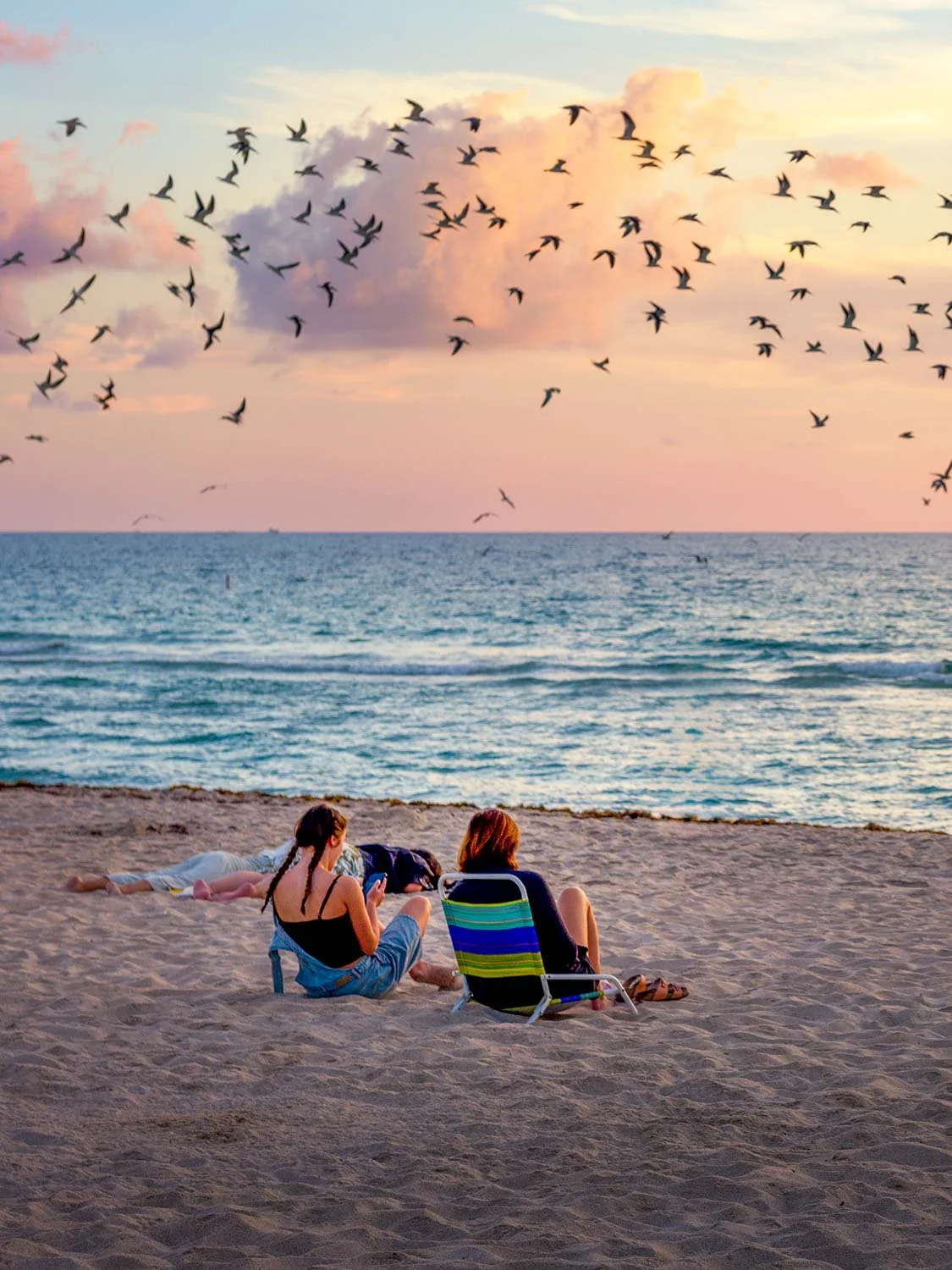 Two women sitting on South Beach sand at sunset watching ocean waves as hundreds of birds flock across a pink sky