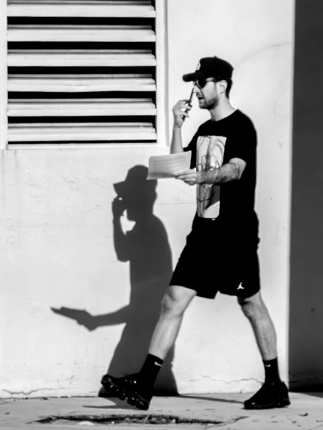 Man on the phone casting a long shadow on a white wall beneath louvered vents in Downtown Miami