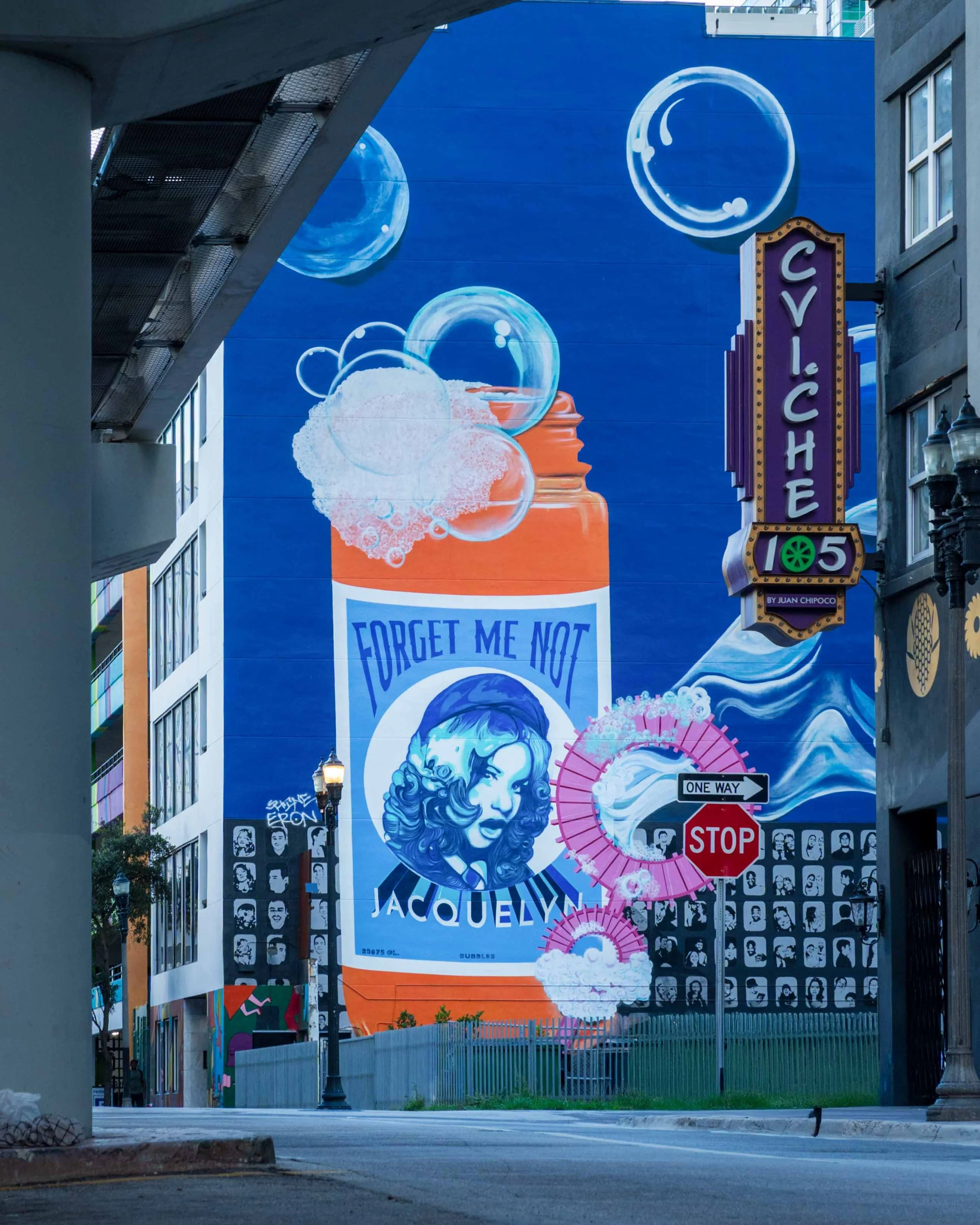 The Forget Me Not bubble bottle on a blue wall, framed from under the elevated track, with the CVI.CHE 105 sign and a stop sign
