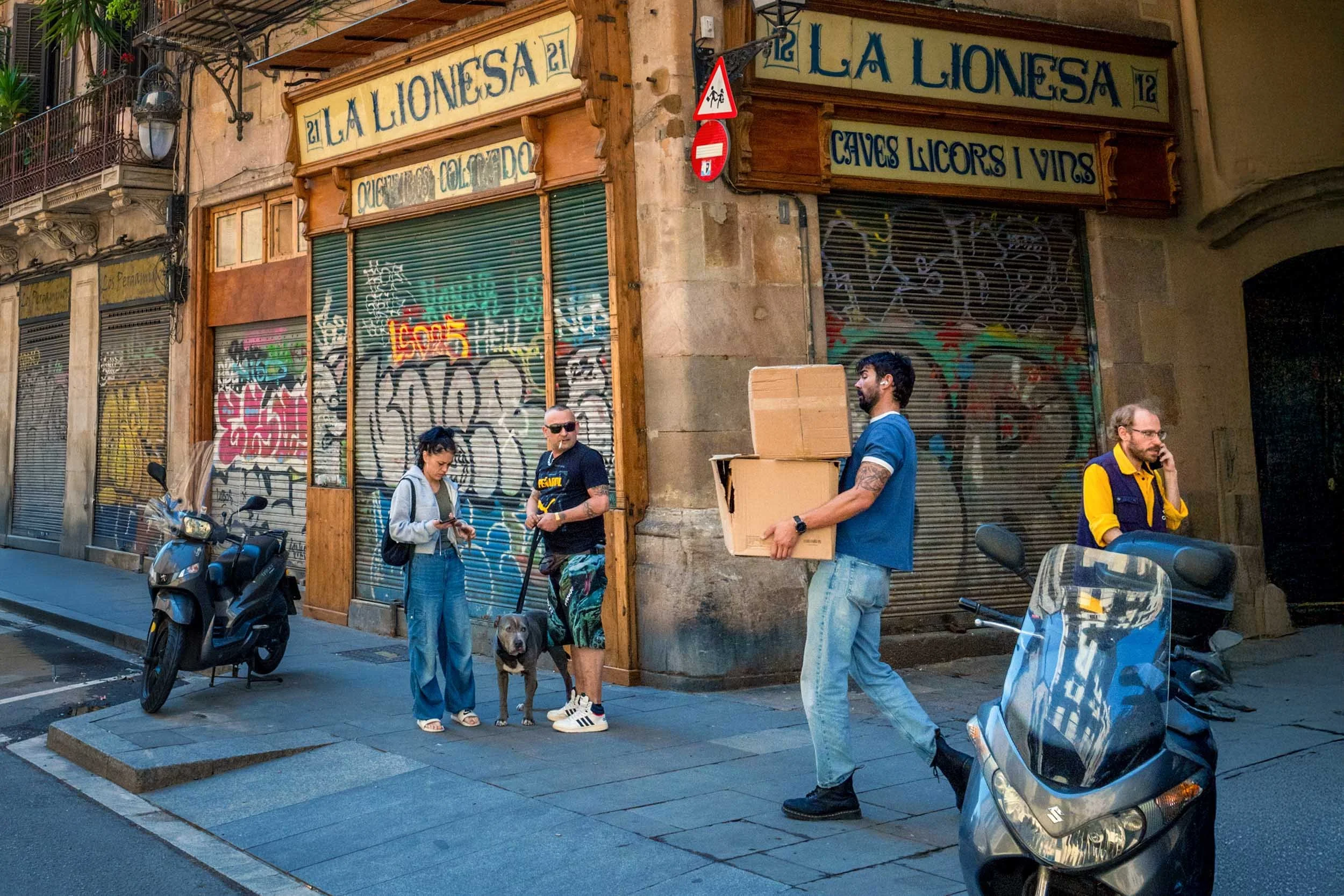 People on a street corner beneath the vintage La Lionesa shop signs and graffiti-covered shutters, a man carrying cardboard boxes past parked scooters