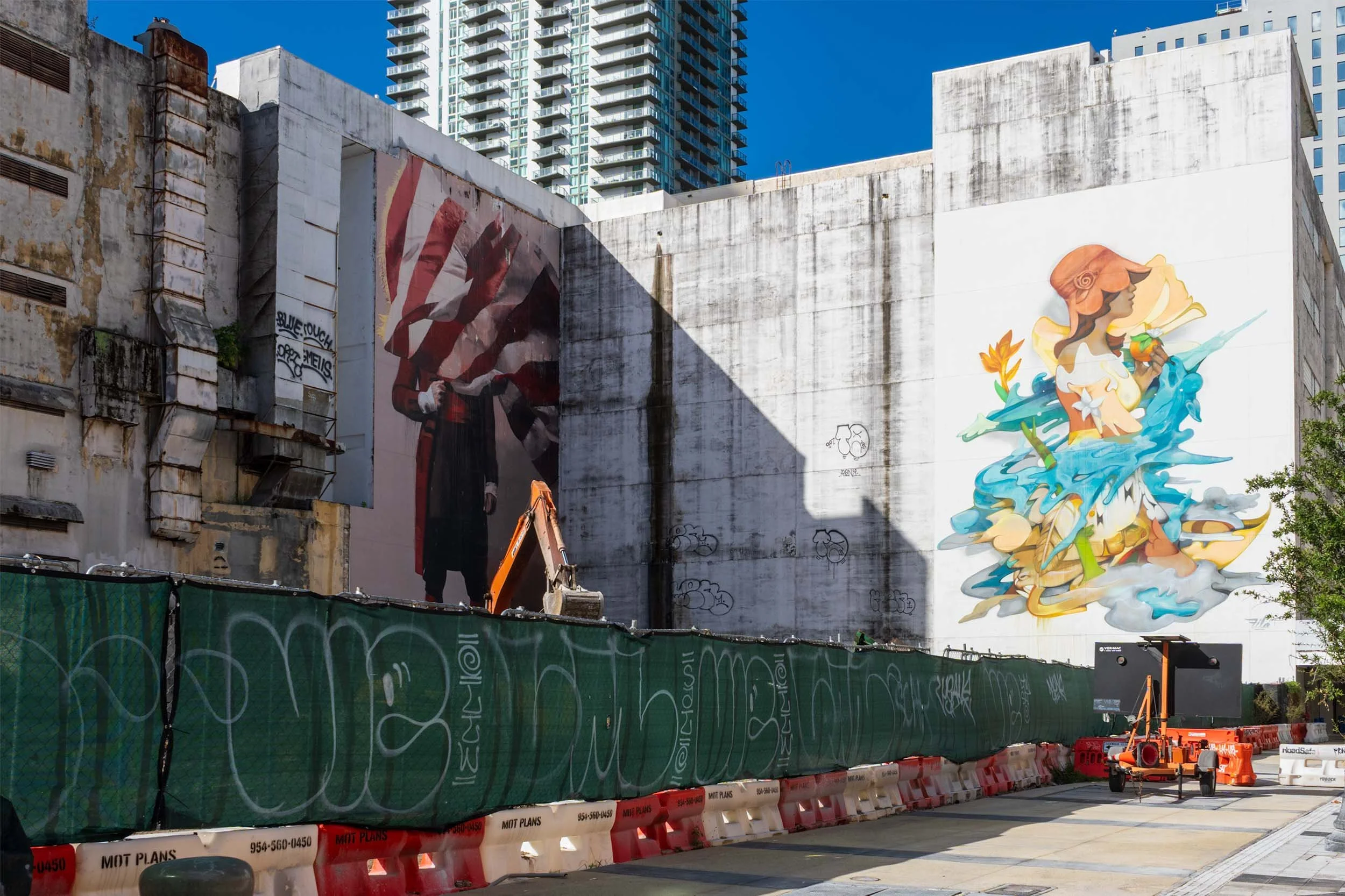 The flag-figure mural with the excavator arm in front of it, framed by leaves
