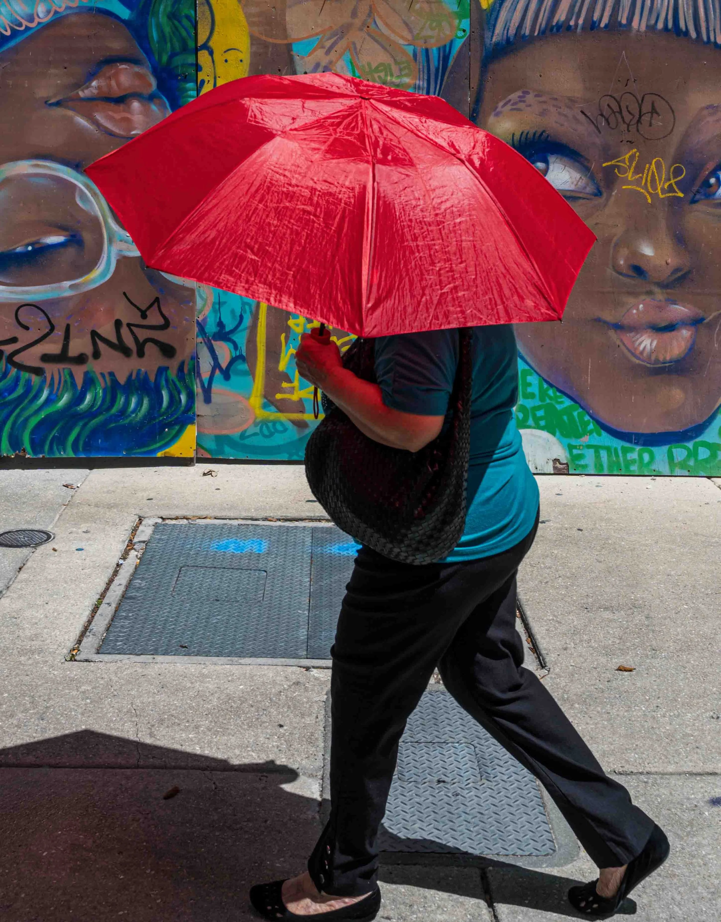 Person walking on urban sidewalk holding bright red umbrella in front of colorful street art mural