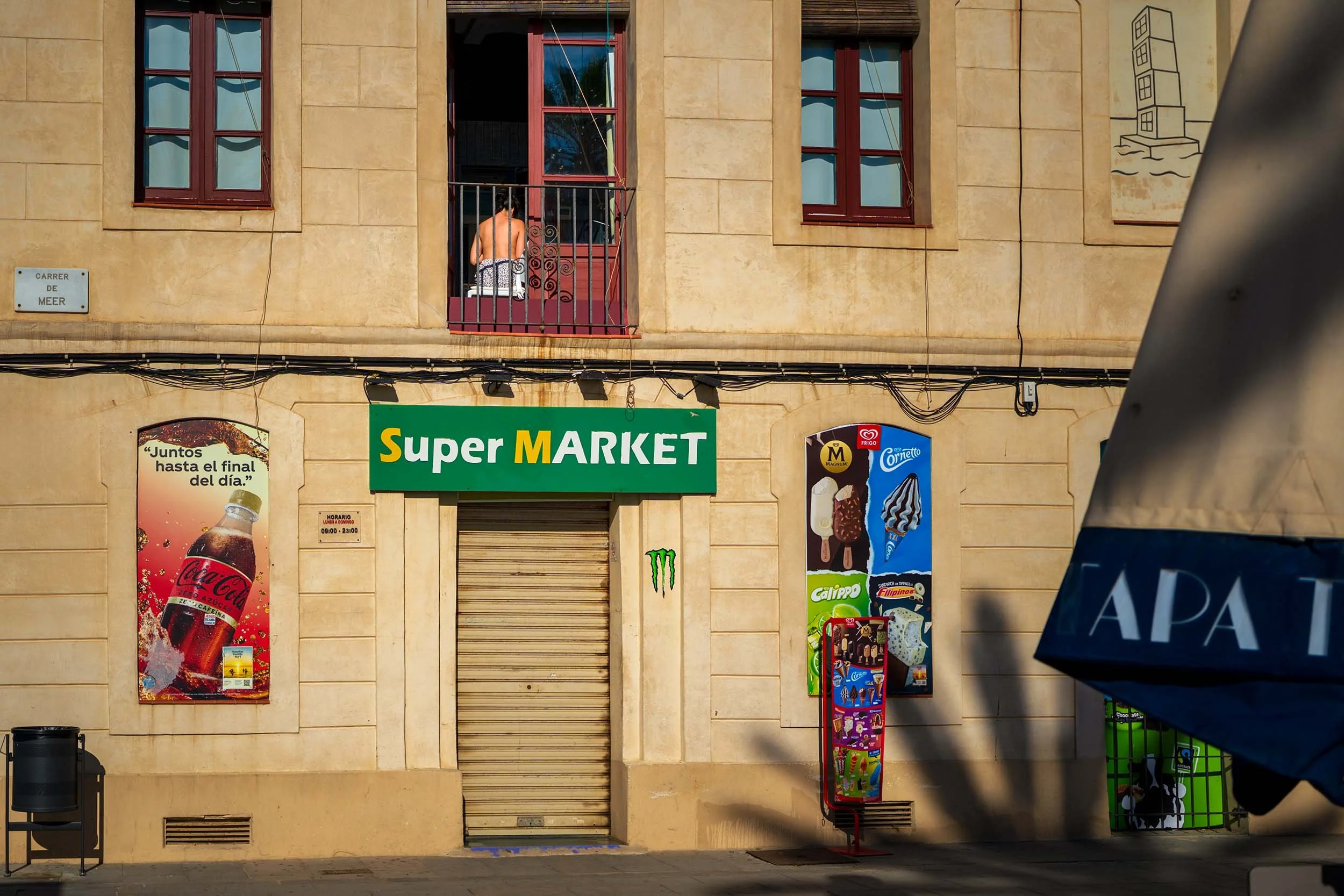 Woman sitting on a balcony above a shuttered supermarket on Carrer de Meer, the facade covered in drink and ice cream