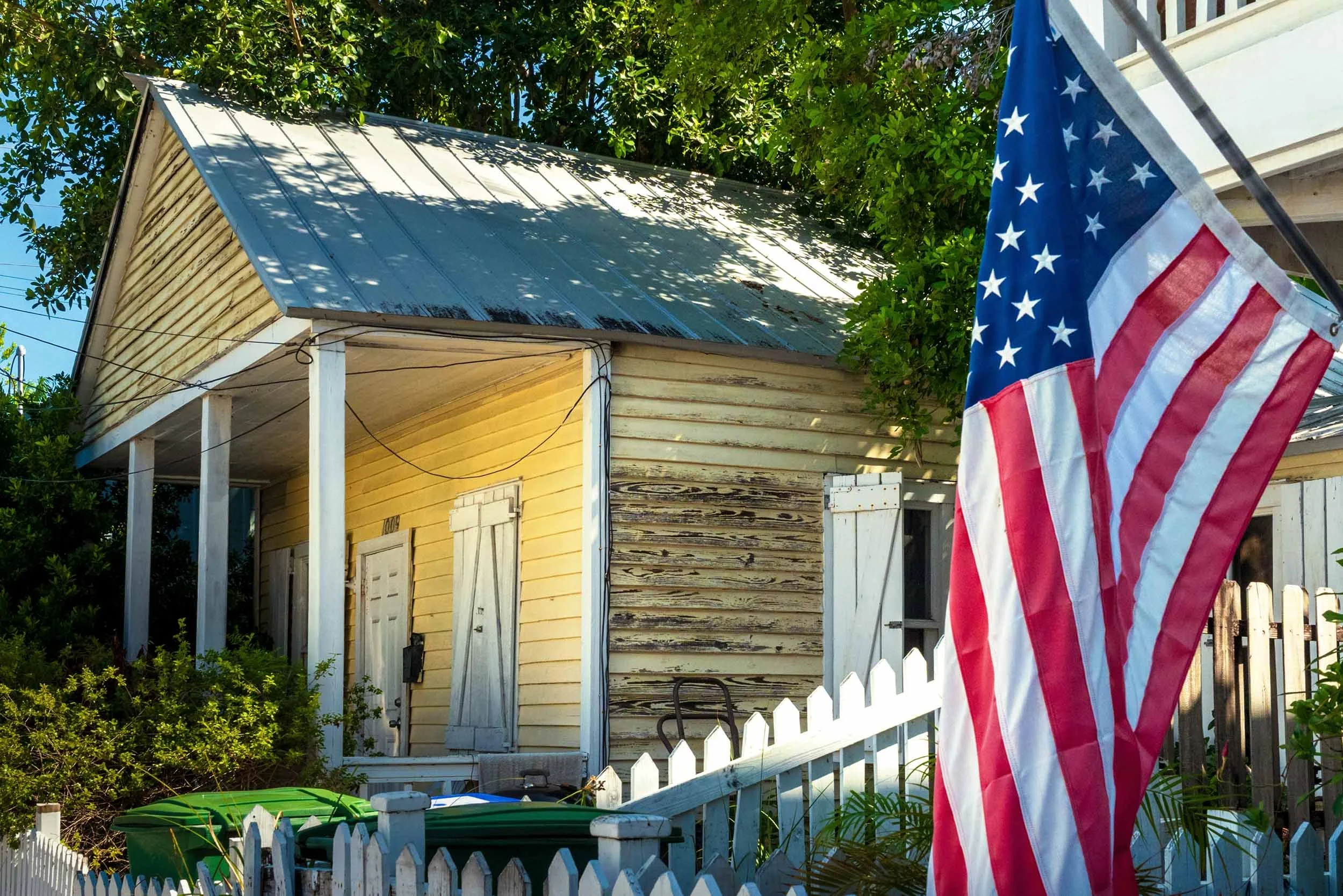 Weathered yellow wood cottage with metal roof and white picket fence beside an American flag in Key West