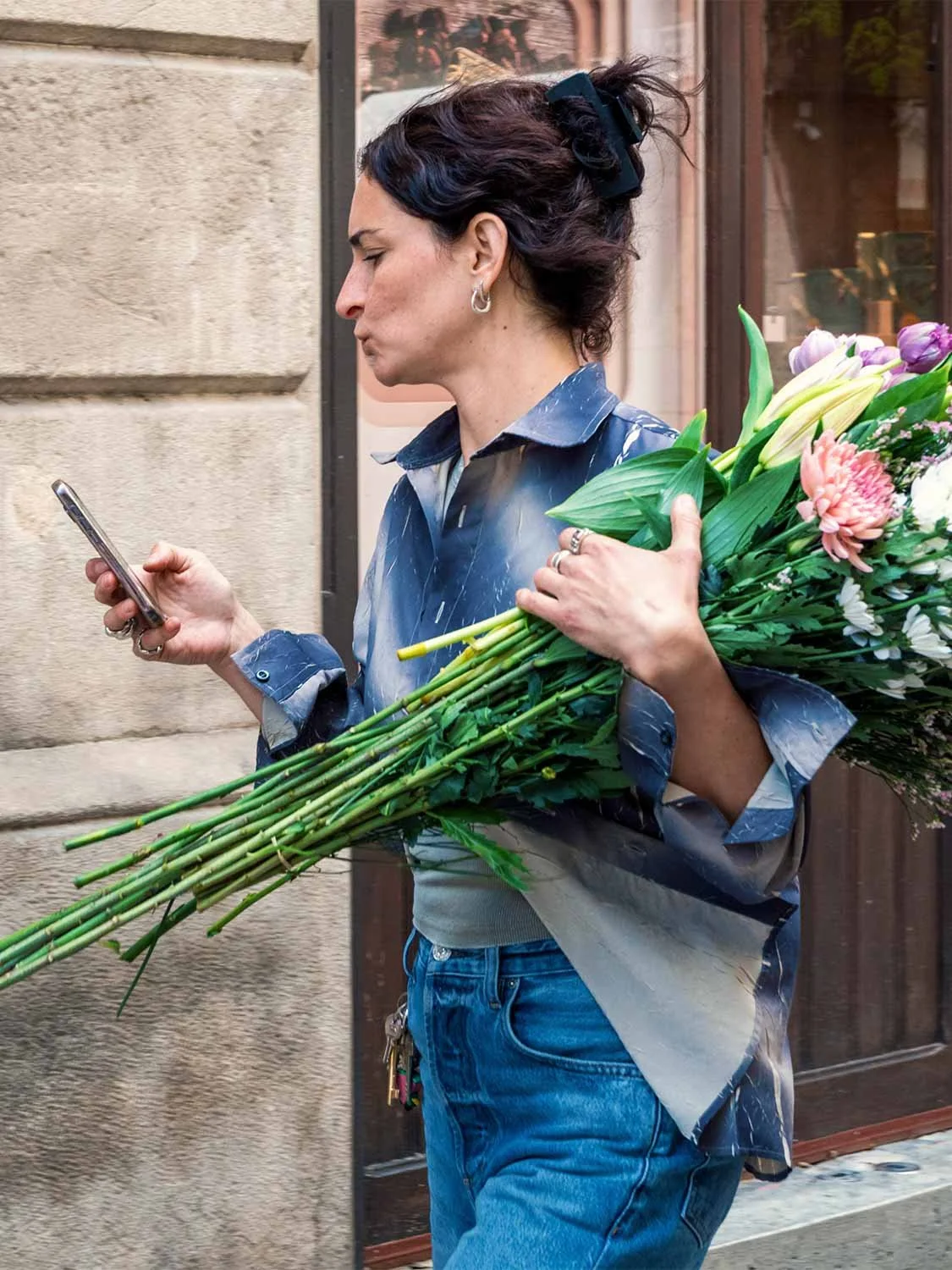 Woman checking her phone while carrying a large bouquet of lilies and tulips past a stone wall in Alfama