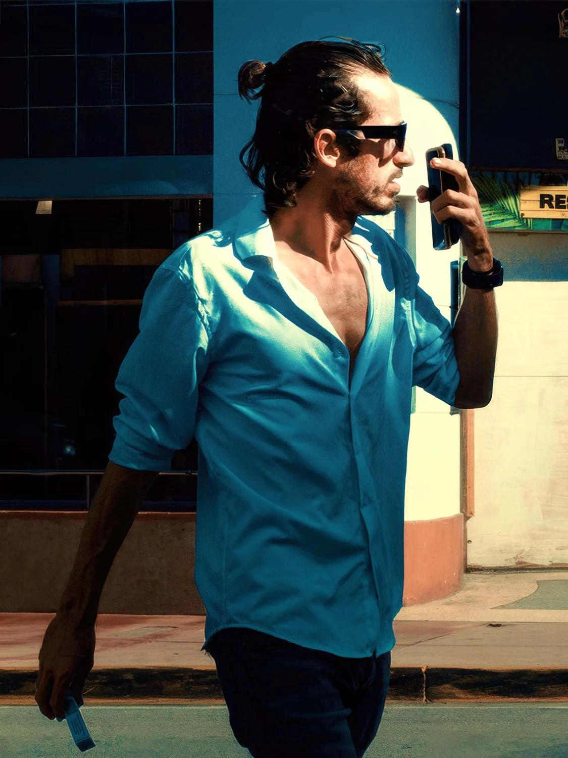 Man in an open teal shirt and sunglasses walking past a South Beach Miami storefront in harsh midday light