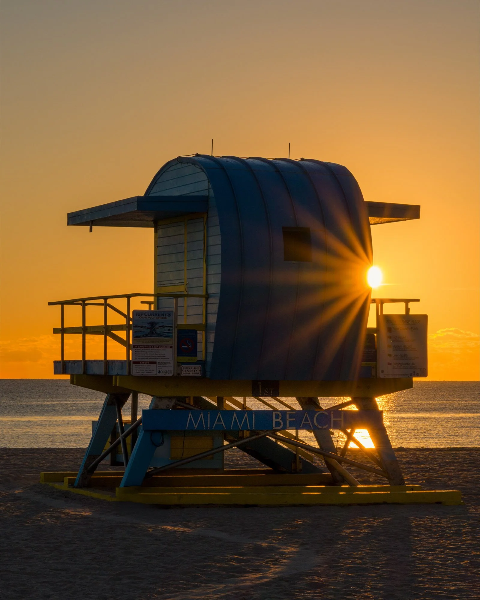 Blue lifeguard stand with a sun flare at its edge under an orange sky, Miami Beach