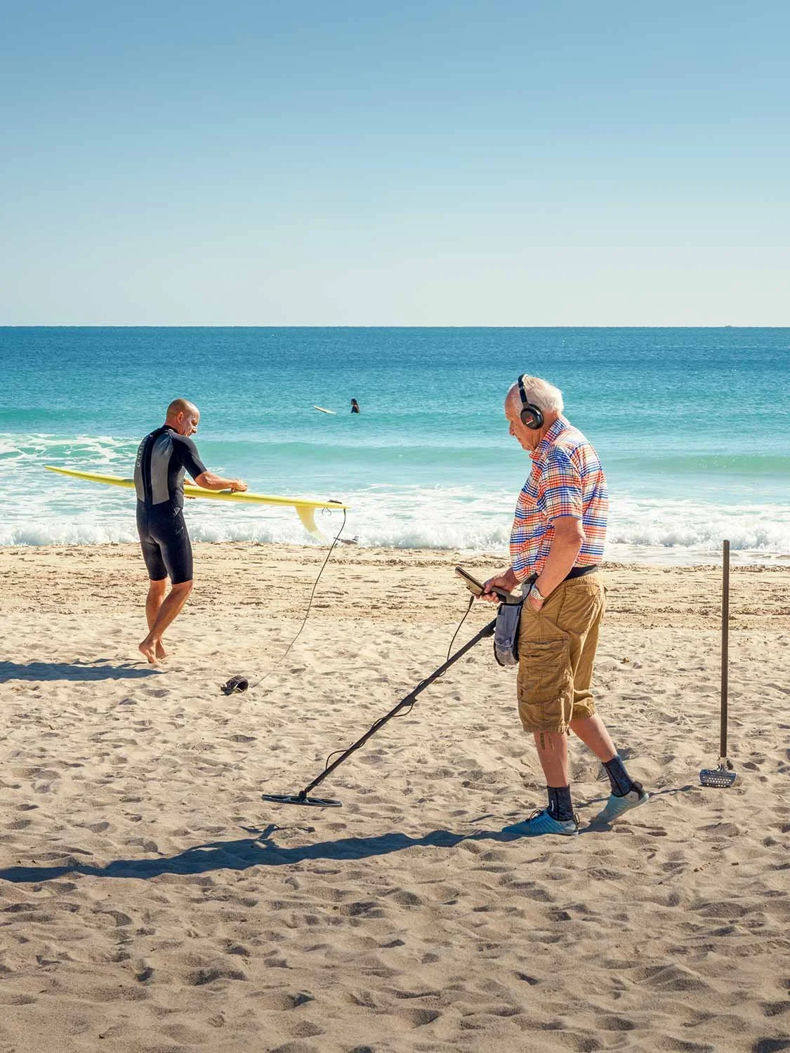 Older man using a metal detector on South Beach sand while a surfer carries a yellow surfboard toward the ocean.