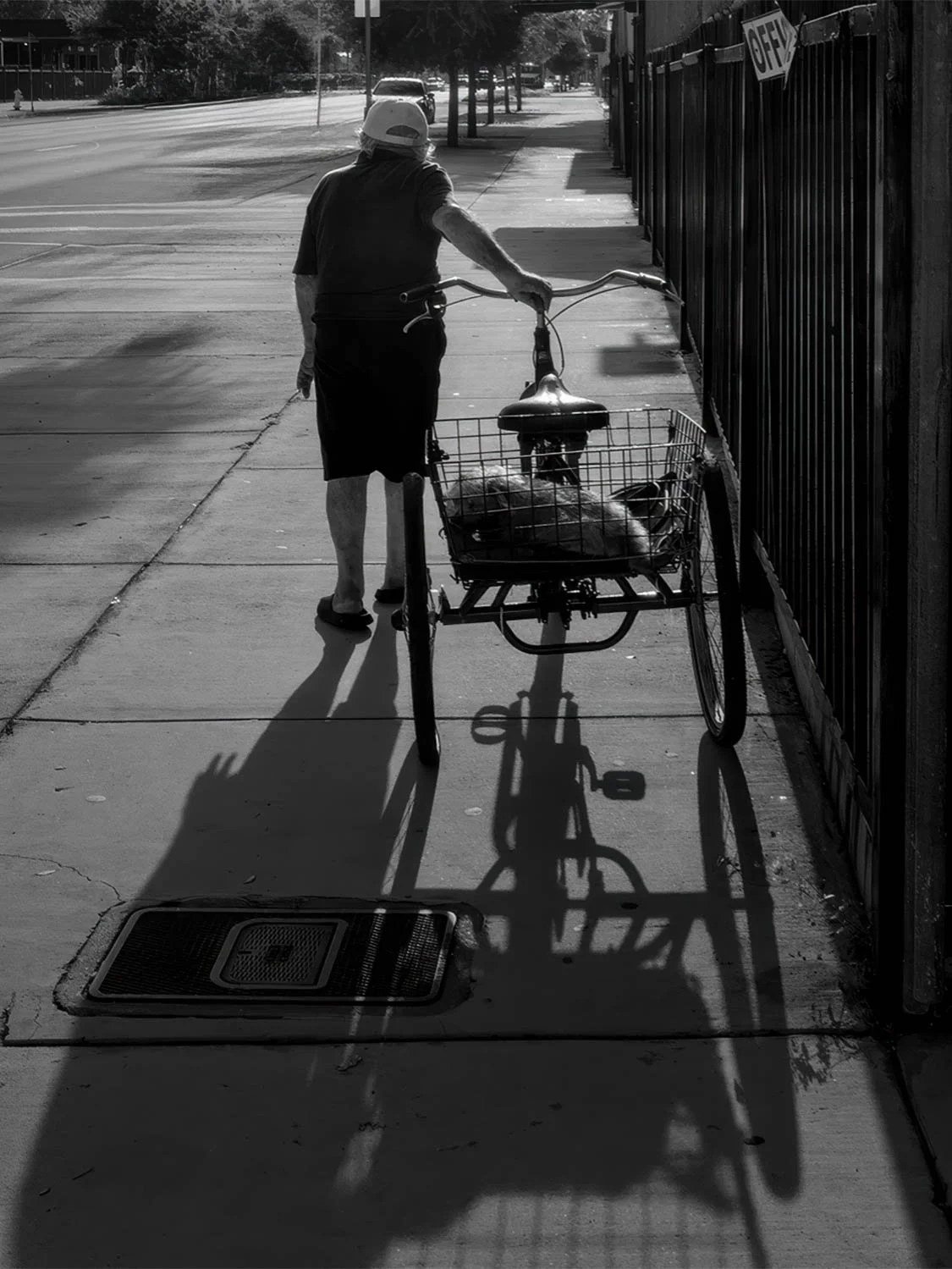 Black and white street photography of elderly person with cargo tricycle basket casting dramatic shadows on city sidewalk.