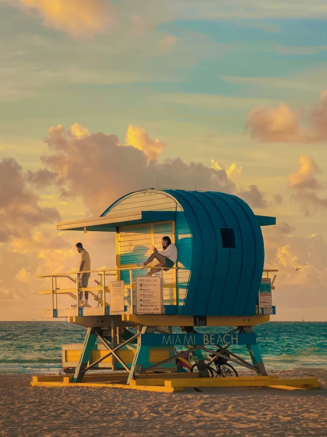 Miami Beach lifeguard tower at 5th Street sunrise with golden sky, teal ocean, and two visitors relaxing.