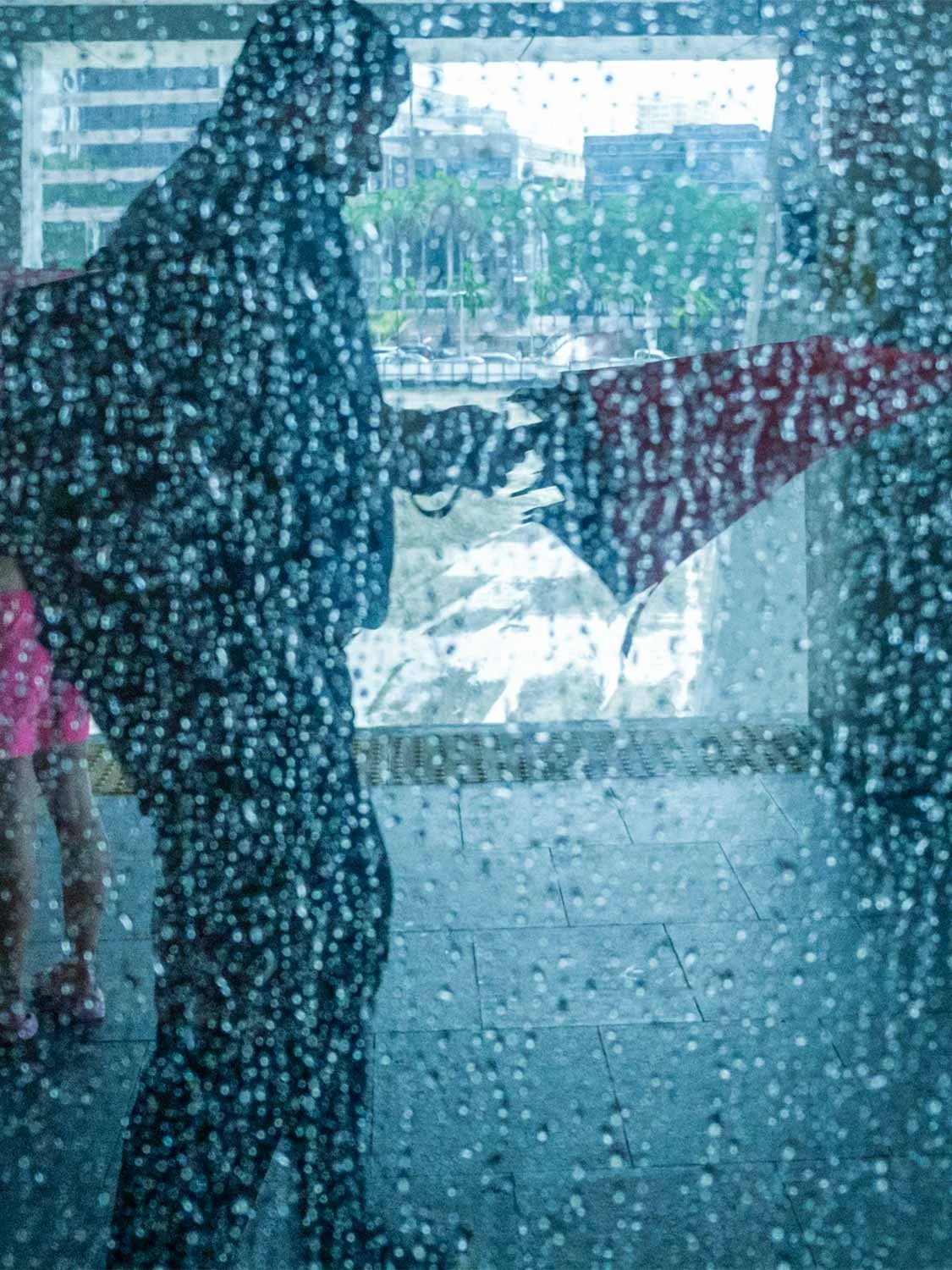Person in hooded raincoat standing at urban entrance, viewed through rain-streaked glass window during heavy rain