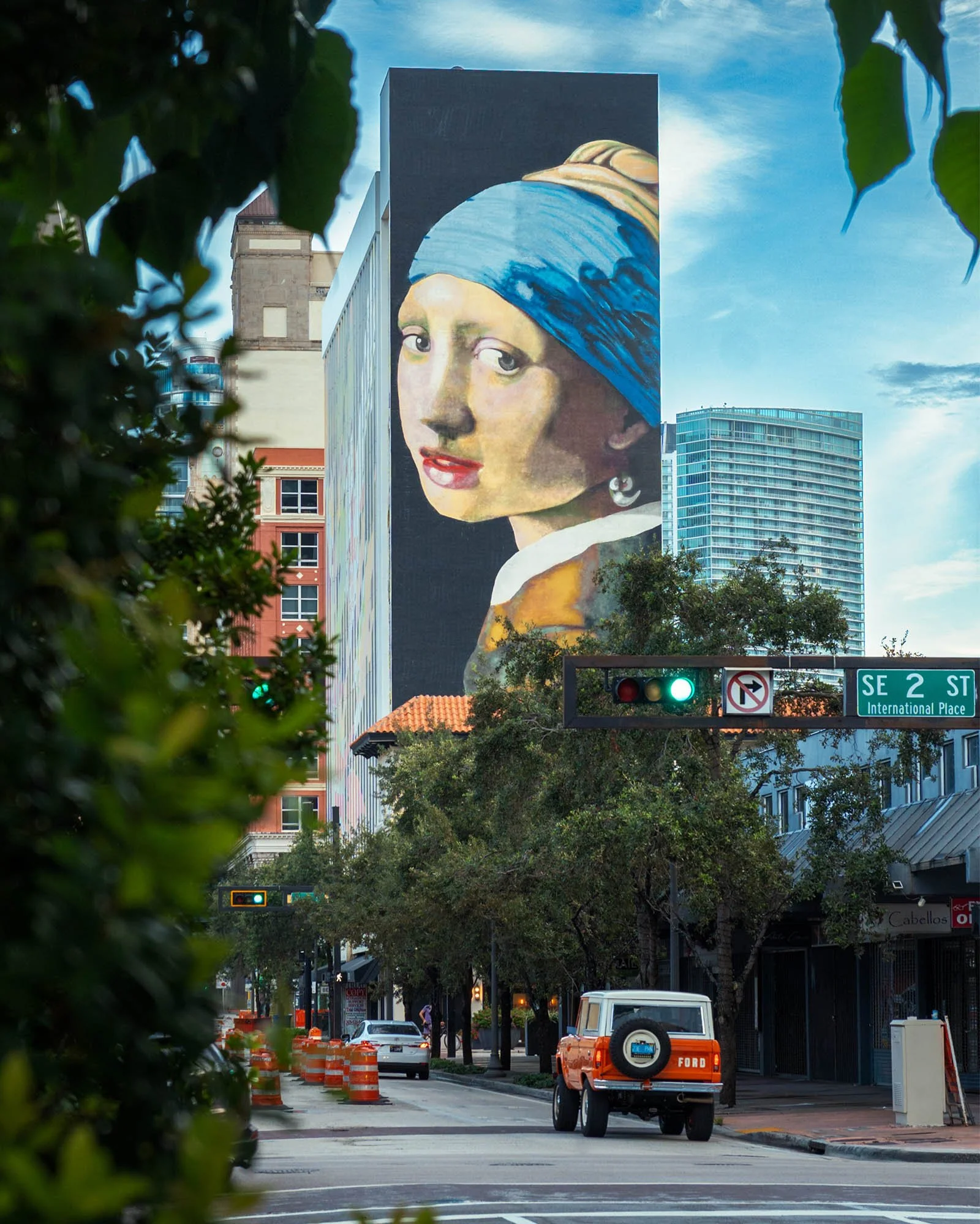 A Girl with a Pearl Earring mural on a tall building, framed by foliage, with the SE 2 St sign and an orange vintage Ford Bronco in the street below