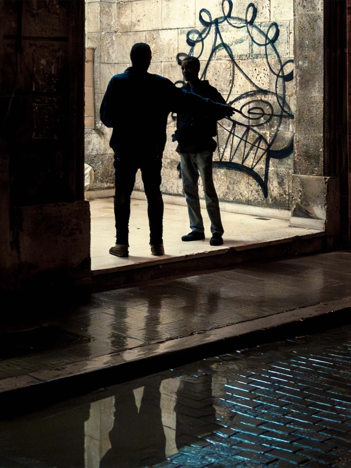 Two men silhouetted in a lit doorway before a graffiti-covered wall, reflected in the wet street in Old Havana