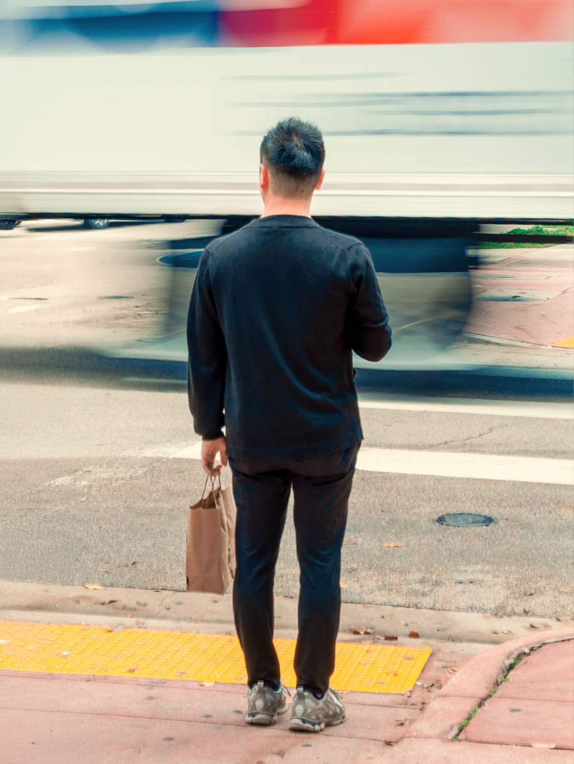 Pedestrian standing at a Miami crosswalk holding a paper bag as a bus streaks past in motion blur