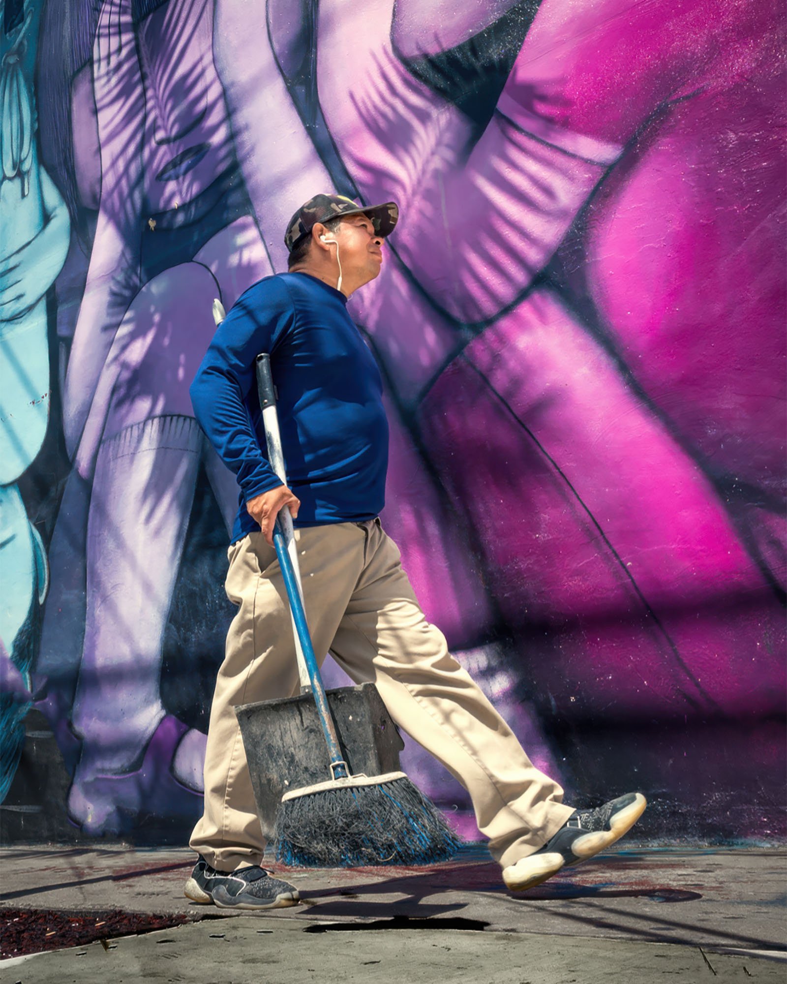 Low-angle shot of a man in a blue shirt and camouflage cap carrying a broom and dustpan past a large purple and pink mural in Wynwood.
