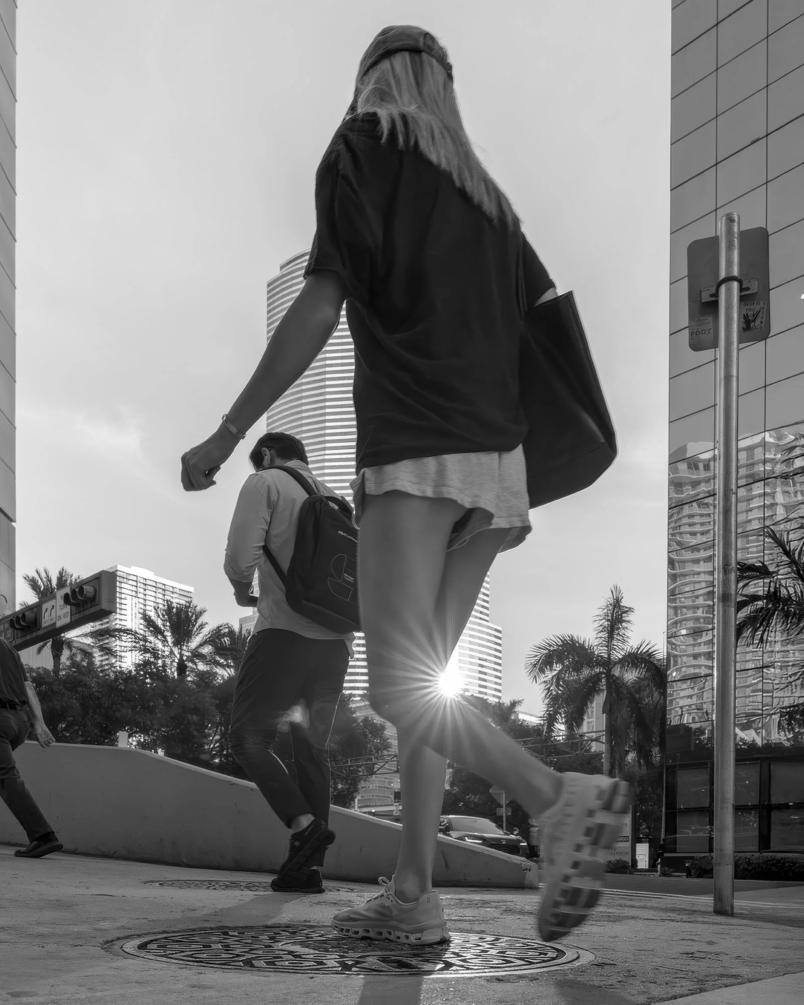 Black and white photo of a woman walking over a manhole cover, sun flare behind her calf, Brickell Ave and Biscayne Blvd Way, Miami