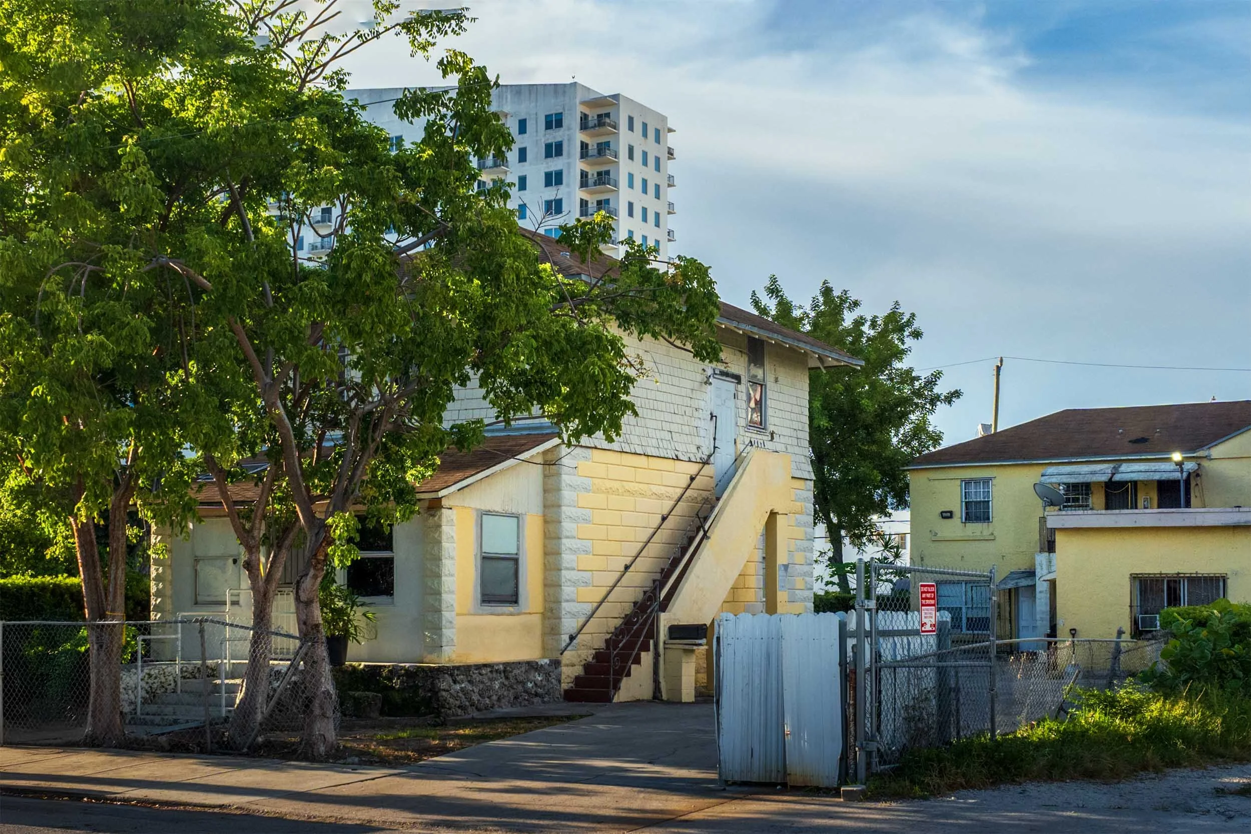 Yellow wood-frame house with an exterior staircase, condo tower behind it, Miami