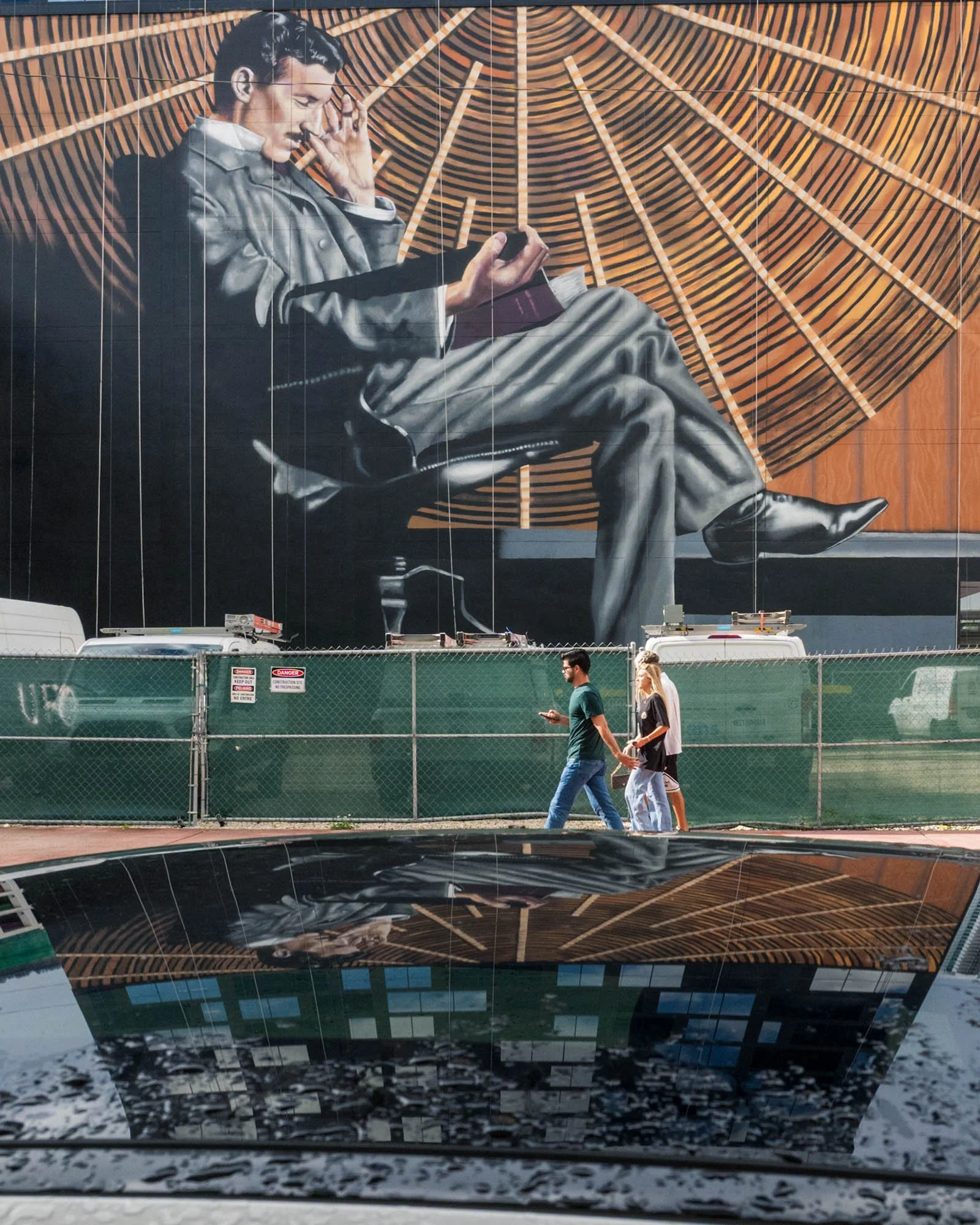 Nikola Tesla seated in a gray suit against wood-toned rays. Pedestrians pass a construction fence below, and the mural reflects in a car hood in the foreground