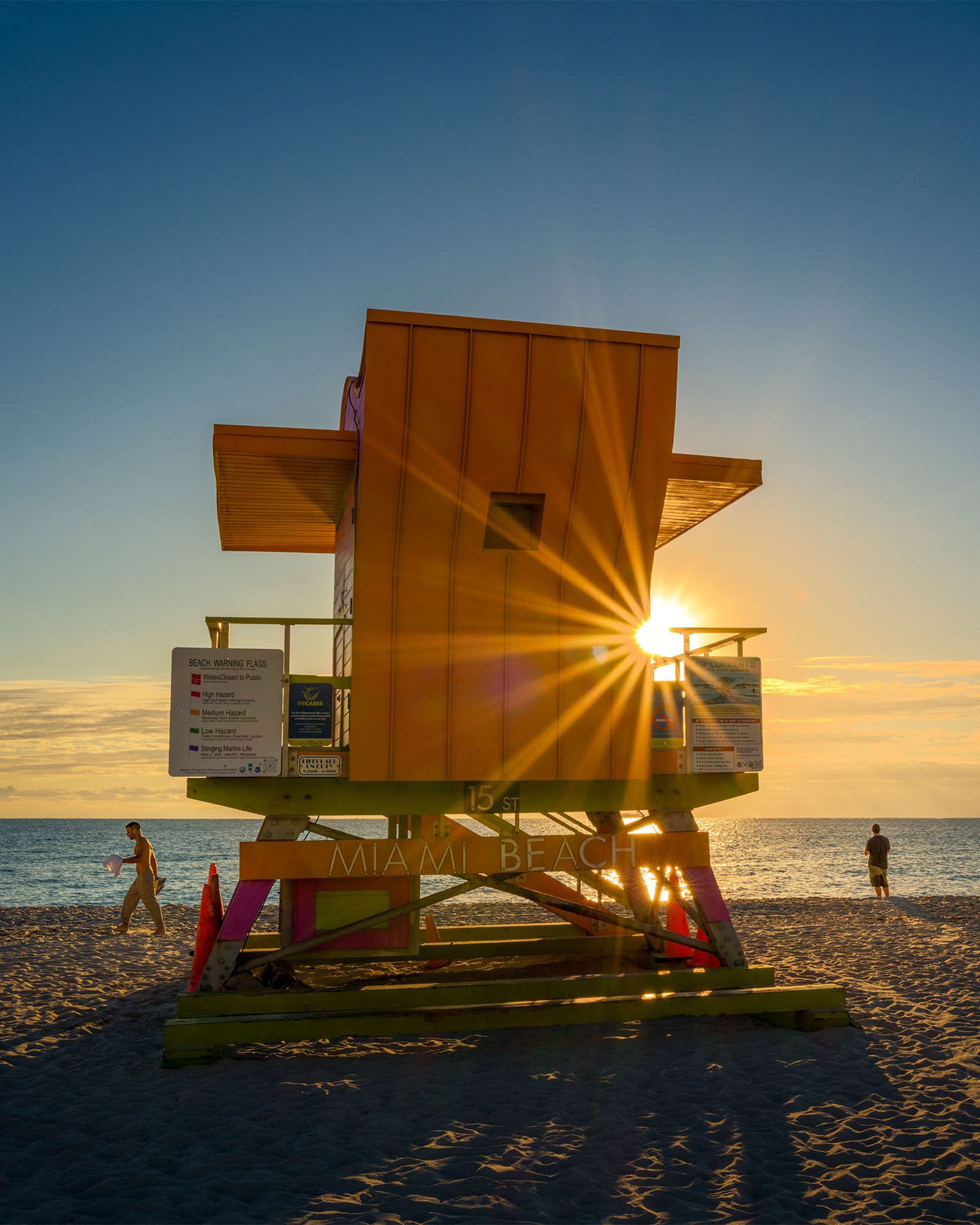 Orange lifeguard stand with a sun flare at the railing and beachgoers on the sand, Miami Beach