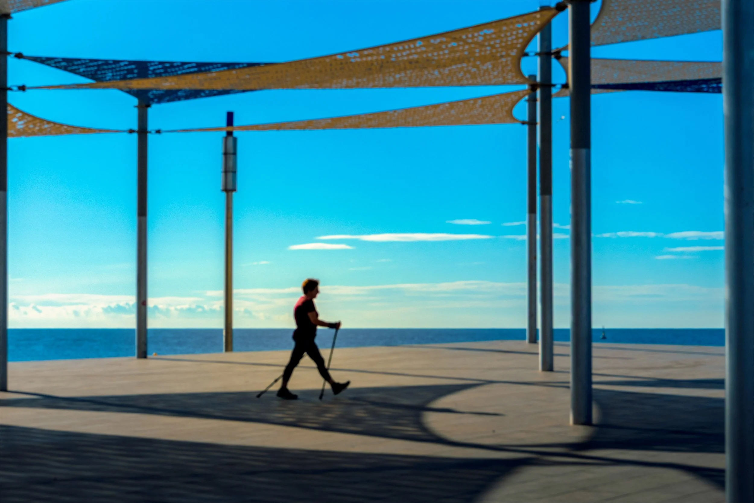 Woman Nordic walking beneath geometric shade sails on a seafront plaza, the Mediterranean on the horizon
