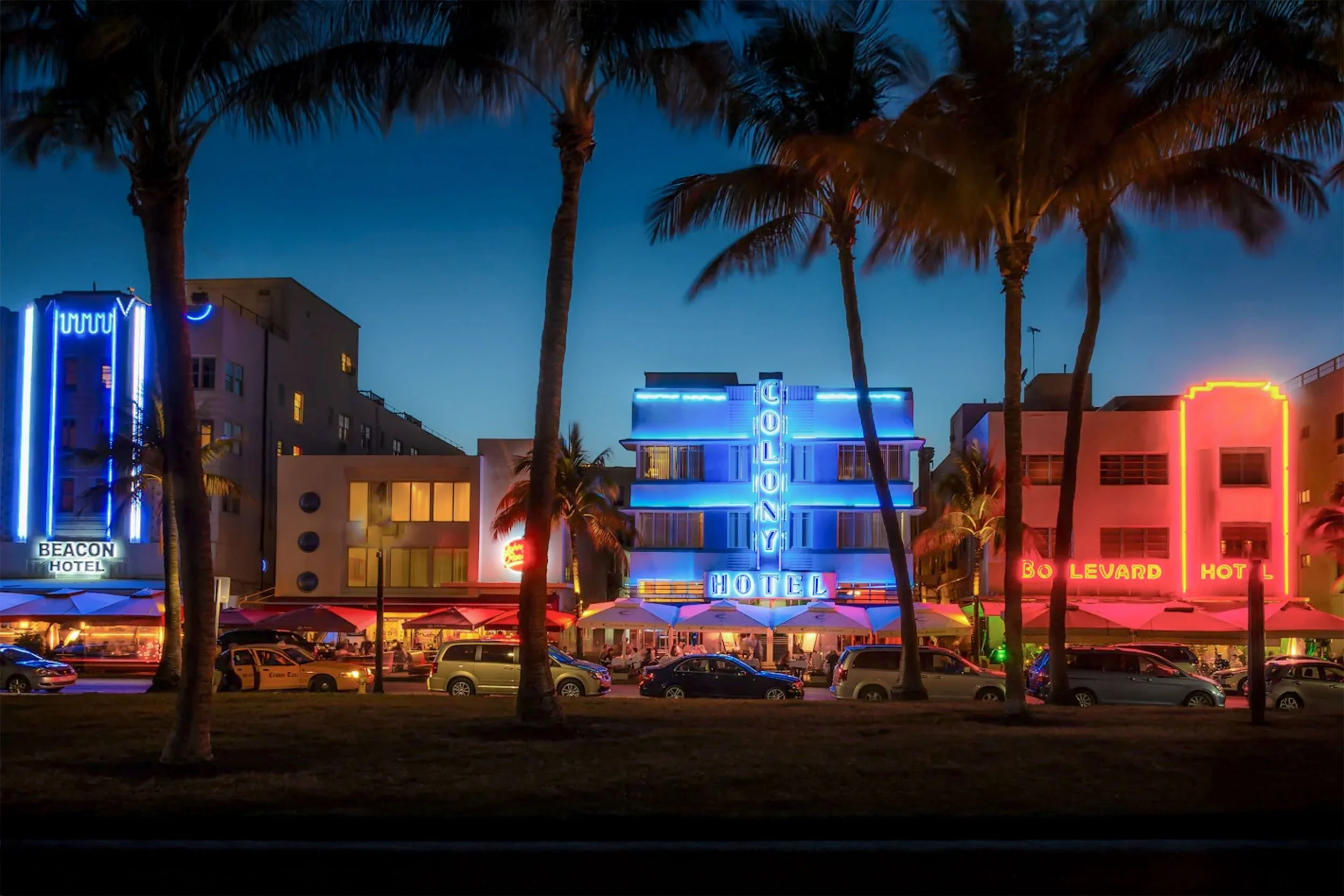Neon-lit Beacon, Colony and Boulevard hotels on Ocean Drive at dusk, framed by palm trees in South Beach