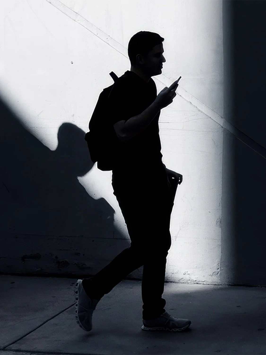 Silhouette of a man with a backpack walking and checking his phone against a bright wall in Barcelona